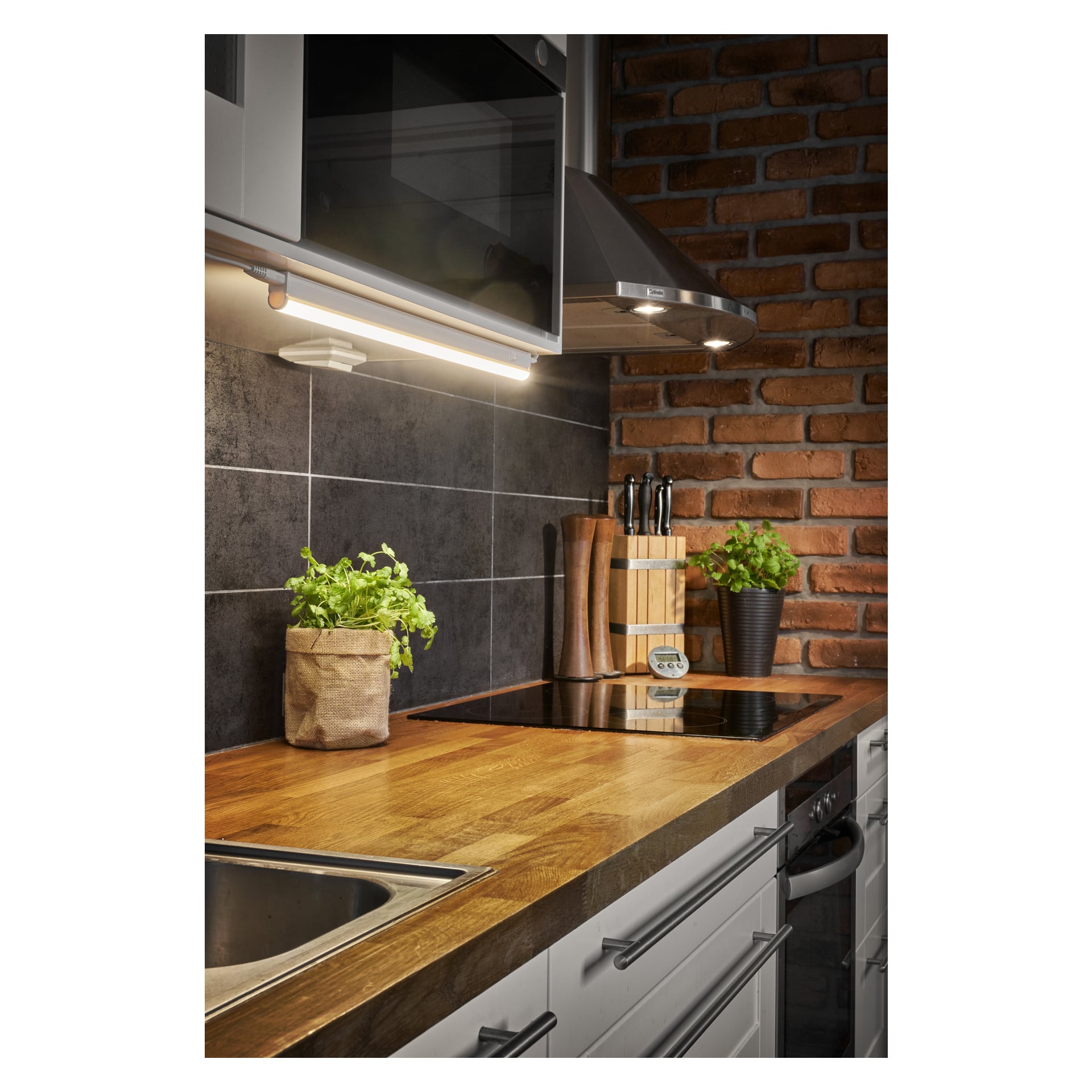Modern kök med träbänkskiva, inbyggd spis, örtplantor, knivblock, salt- och pepparkvarnar, diskho i rostfritt stål, tegel- och kakelskydd samt Anslut Worktop LED-belysning för belysning under skåpen.