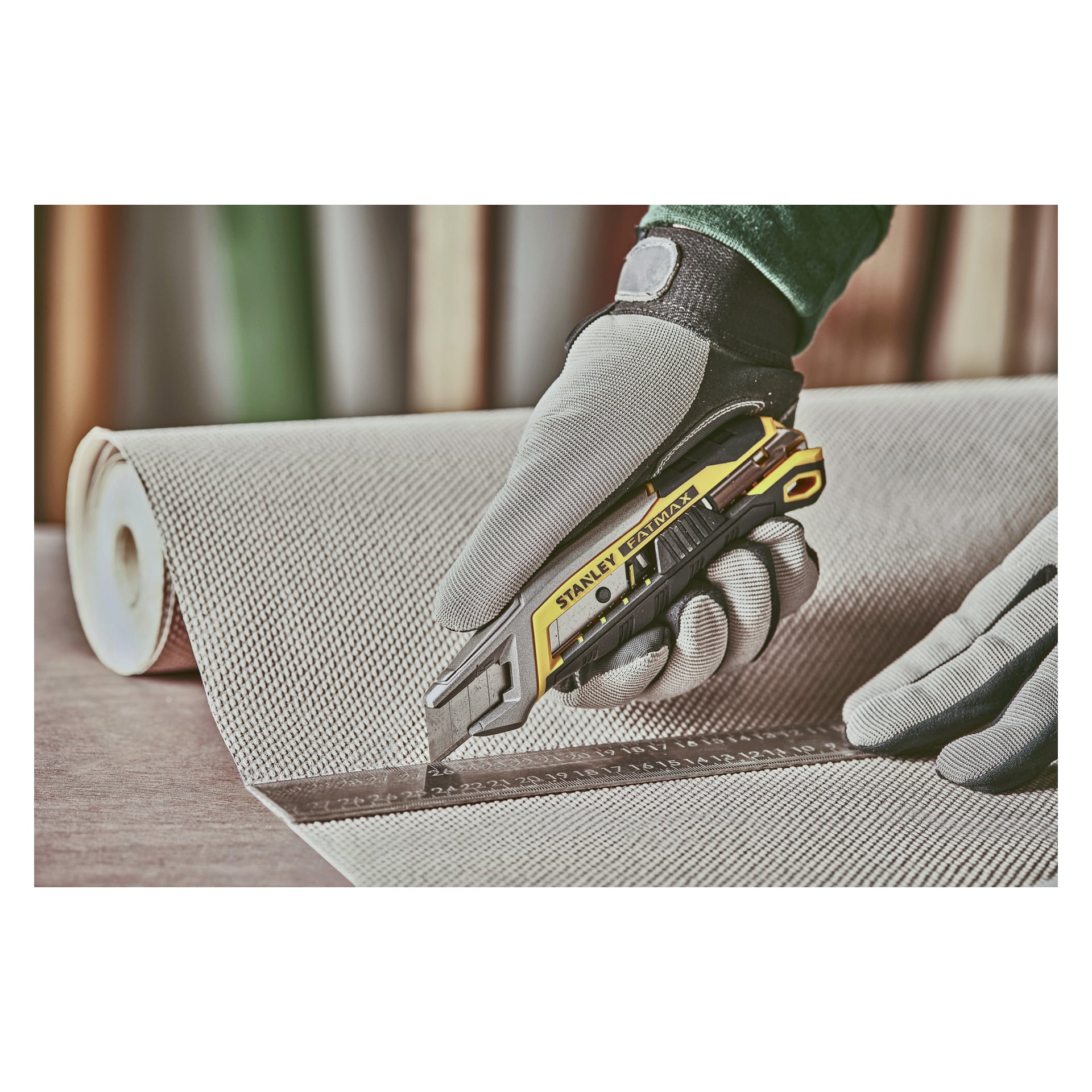 En handskbeklädd hand klipper strukturerad tapet som ligger ut platt, med en Stanley Fatmax 18 mm avbrytningskniv och en metalllinjal med centimetermarkeringar.
