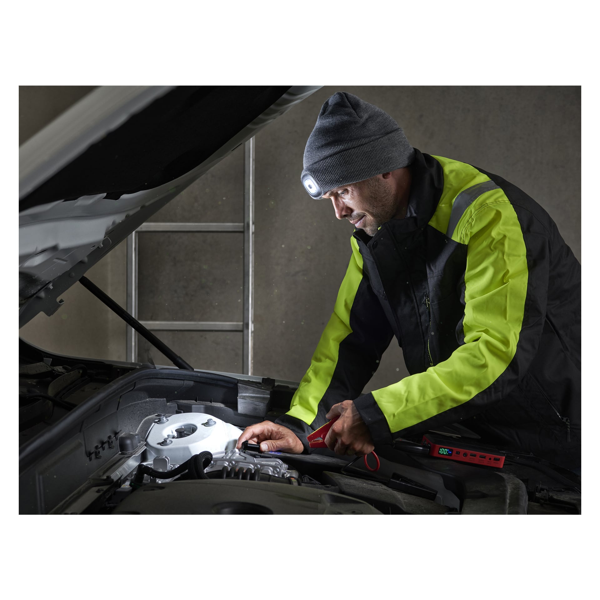 Mit einer neongelben Jacke und einer Kayoba Outdoor-Mütze mit LED-Licht schaut eine Person unter die Motorhaube eines Autos, hält sowohl ein rotes Werkzeug als auch ein tragbares Diagnosegerät in der Hand. Im Hintergrund steht eine Metallleiter.