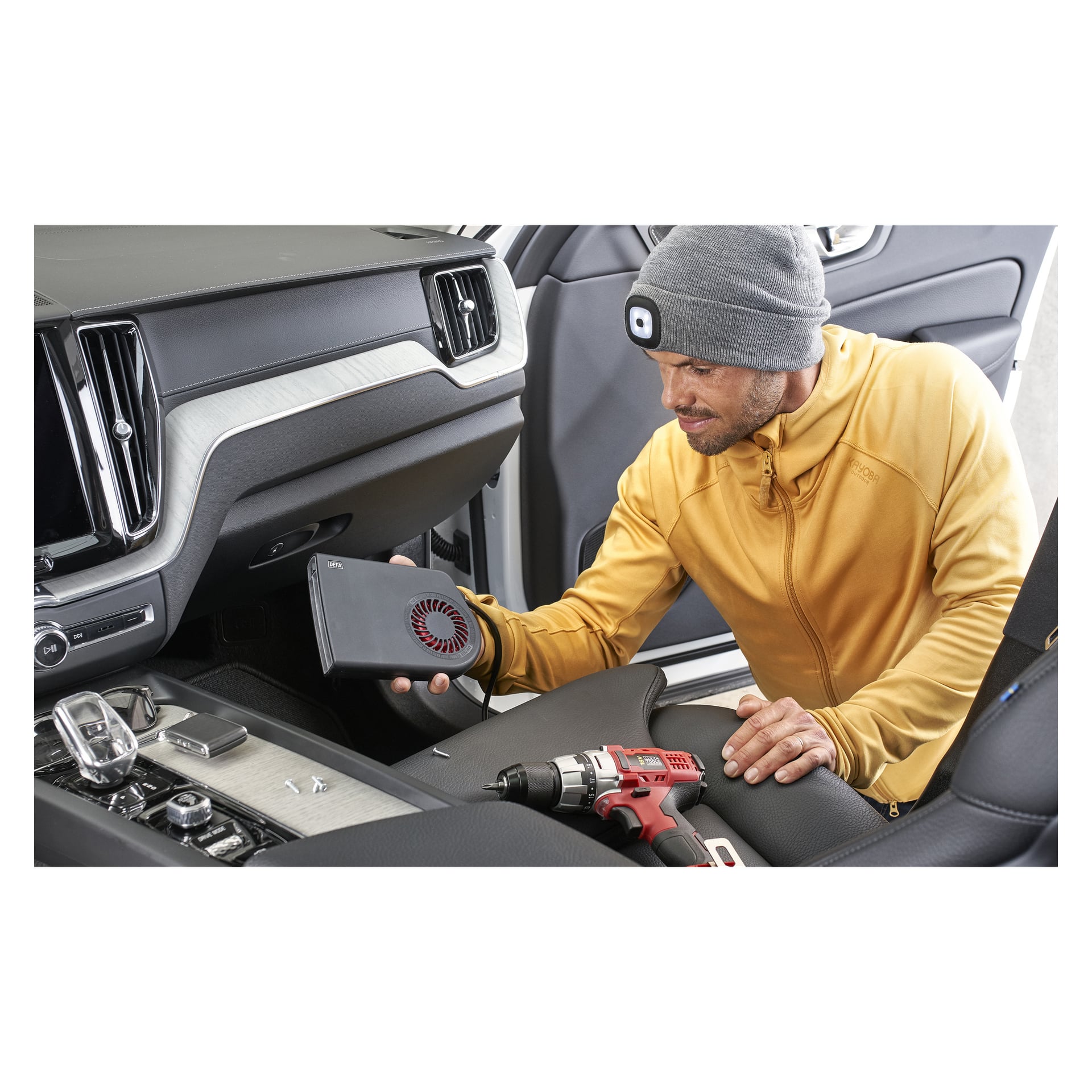 In einer gelben Jacke und einer Kayoba Outdoor-Mütze mit LED-Licht untersucht eine Person das Handschuhfach des Autos, das ein kleines, belüftetes schwarzes Gerät hält. Ein Bohrer wird auf dem Sitz neben ihnen platziert.
