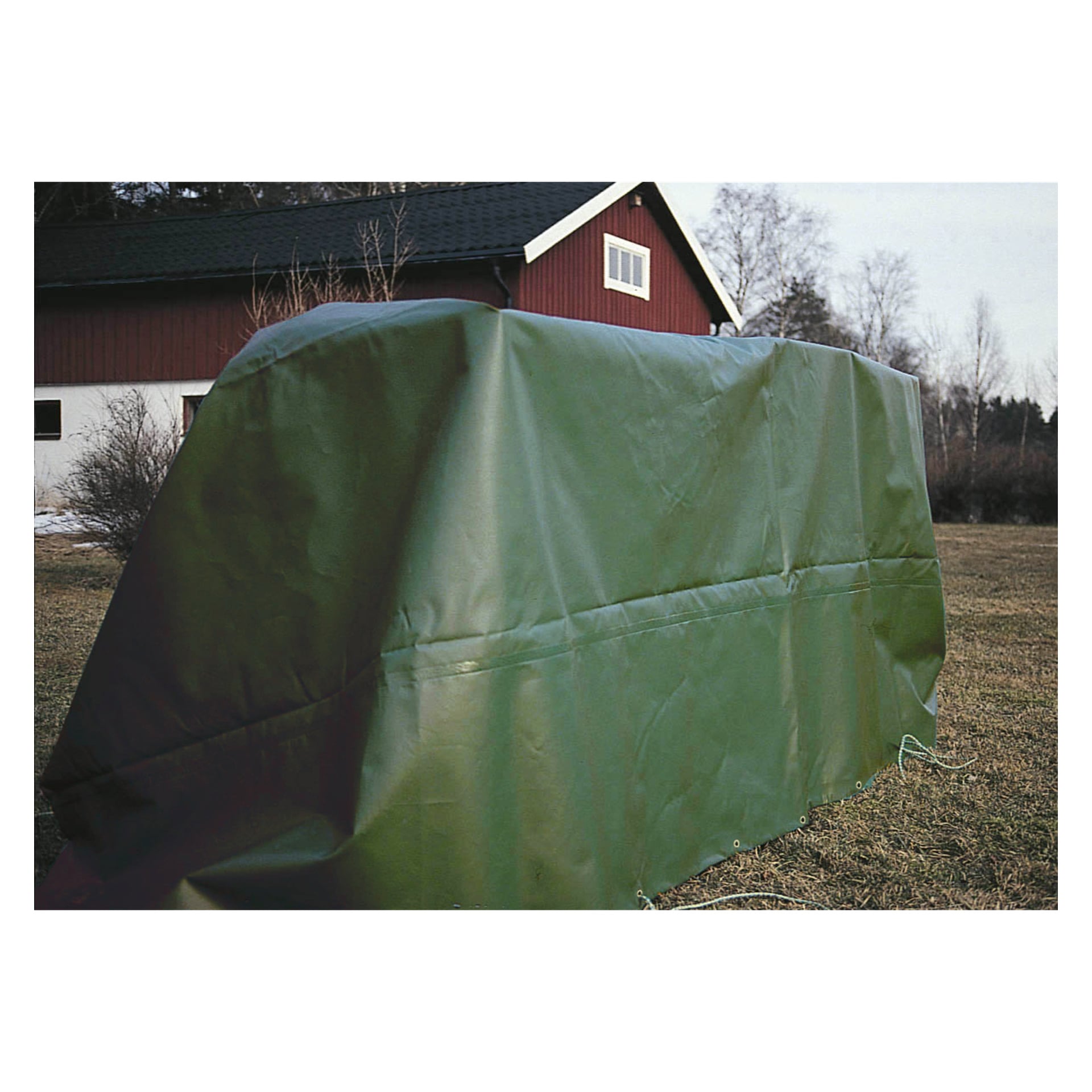 En stor grön Hard Head-presenning är draperad över ett okänt föremål på en gräsbevuxen gård, med en röd lada-liknande struktur och kala träd i fjärran.