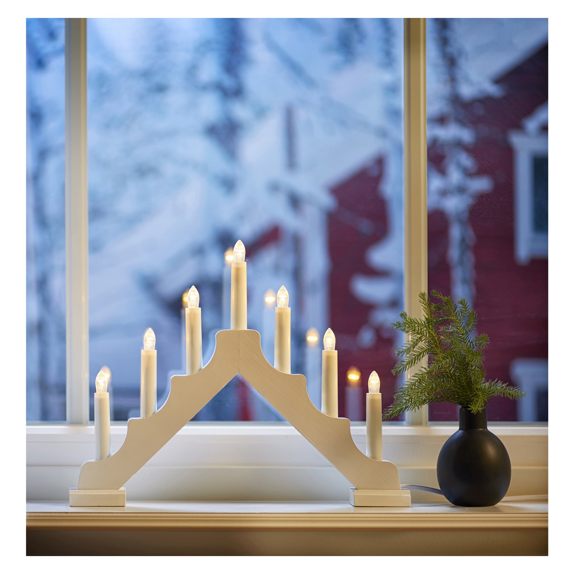 En EKVIP LED-adventsljusstake med sju tända vita ljus står på en fönsterbräda bredvid en liten grön växt i en svart vas. Utanför fönstret finns snötäckta träd och ett rött hus.