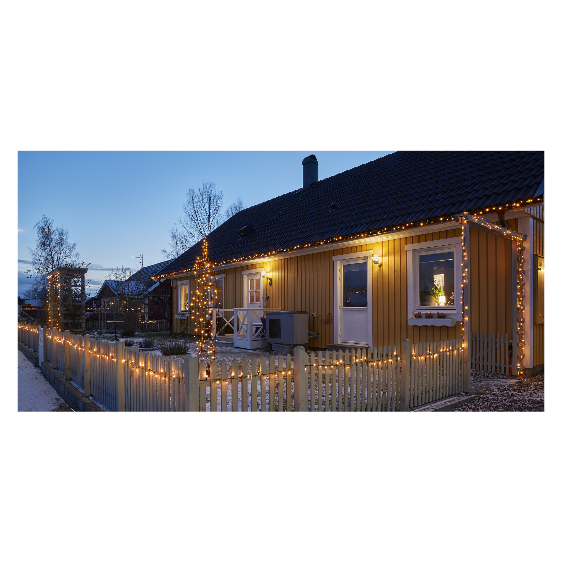 Bei Einbruch der Dämmerung leuchtet ein gelbes Haus mit weißen Leisten unter EKVIP-LED-Lichterketten, die das Dach und den Zaun zeichnen, mit einem kleinen Lattenzaun im Garten, einem beleuchteten Baum und einem leichten Schneestaub auf dem Boden.
