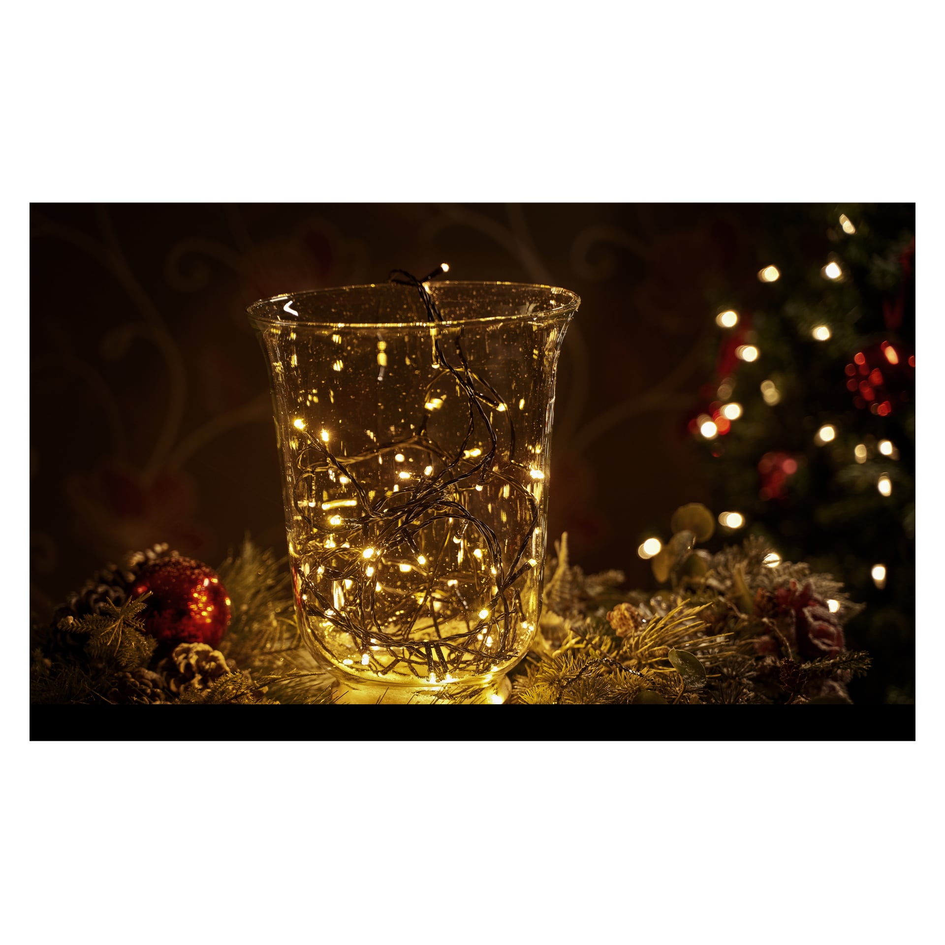 Ein Arrangement zeigt eine klare Glasvase, gefüllt mit EKVIP LED-Lichterketten, umgeben von Tannenzapfen, Grünzeug und Ornamenten auf einer festlichen Oberfläche. Im verschwommenen Hintergrund steht ein Weihnachtsbaum, geschmückt mit roten Ornamenten und Lichtern.