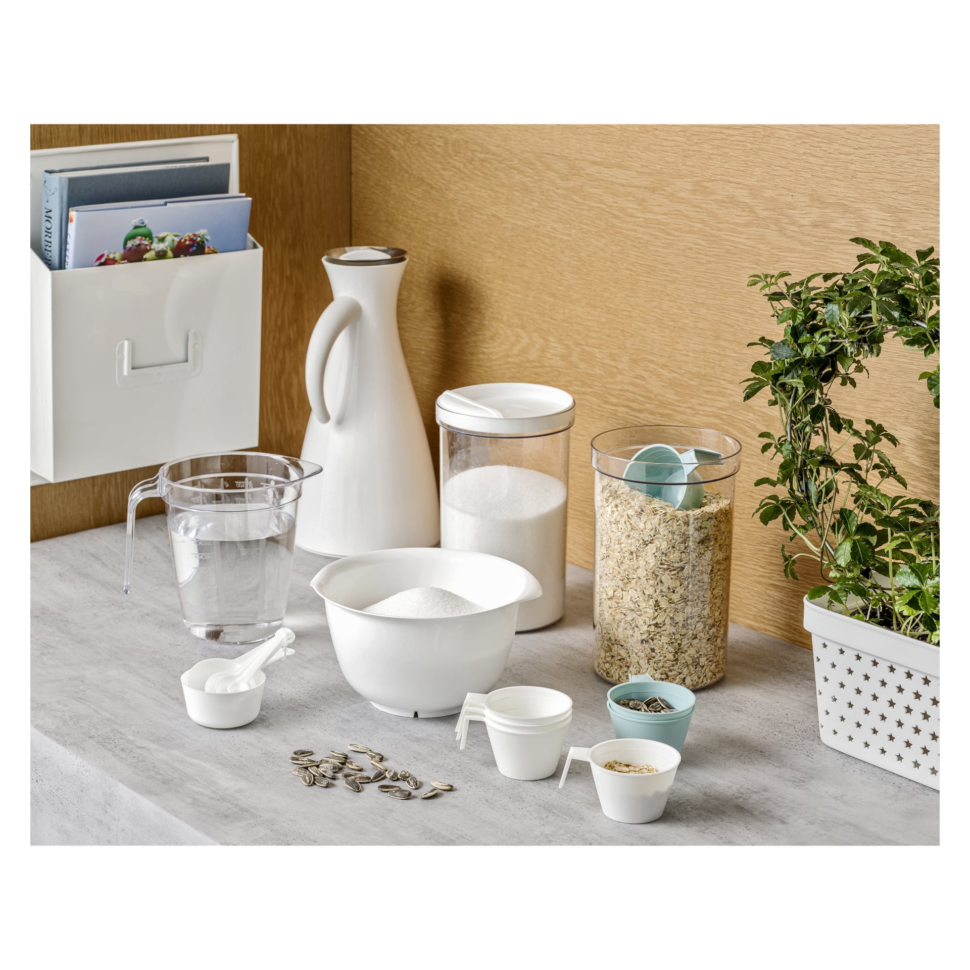 Keittiötasolla on kannu, kulhollinen sokeria, kaurapurkki kauhalla, toinen sokerirasia, kolme Menuett Decilitre -annosta vedellä ja auringonkukansiemenillä, mittalusikat sekä ruukkukasvi.