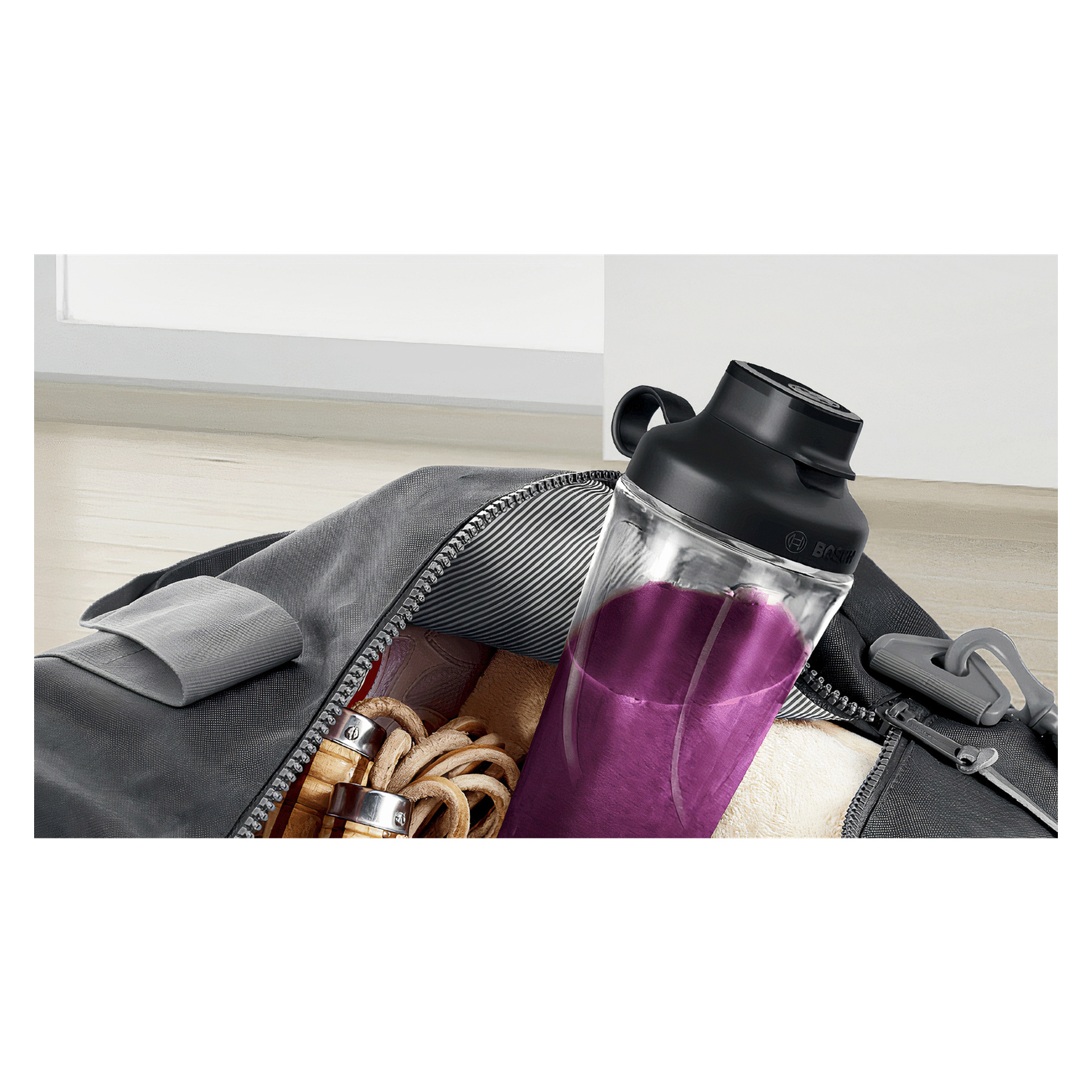 En Bosch Home Blender MMB2111S shakerflaska med svart lock, fylld med lila vätska, finns delvis i en öppen grå gymväska. Bredvid flaskan ligger en handduk och ett hopprep. Väskan vilar på trägolvet inomhus.