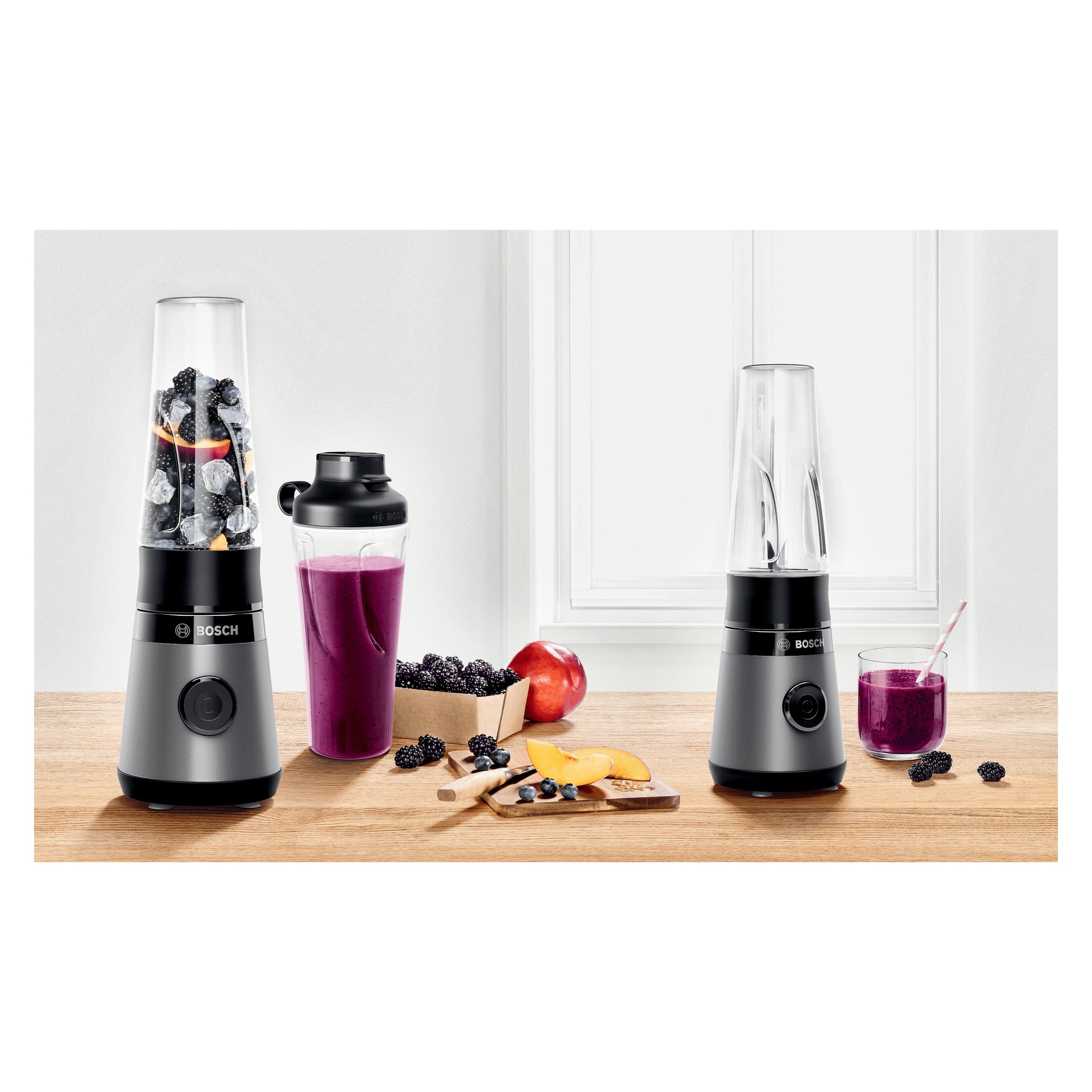 Bosch Home Blender MMB2111S står på en träbänk vid ett fönster, fylld med frukt och is. Bredvid den står en shakerflaska med en lila smoothie, strödda bär, en persika och ett glas med en färdig smoothie.