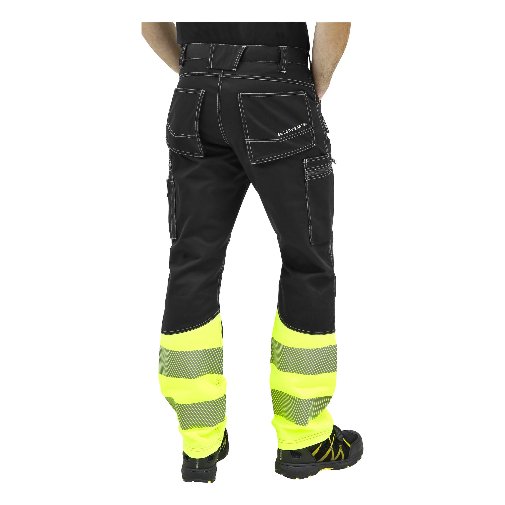 Eine Person steht mit den Armen an den Seiten, von hinten gesehen, und trägt Bluewear-Hochsichtbarkeitshosen ohne Nageltaschen. Die Hose hat reflektierende gelbe Abschnitte, weiße Nähte, mehrere Taschen und die Person trägt schwarze Schuhe.