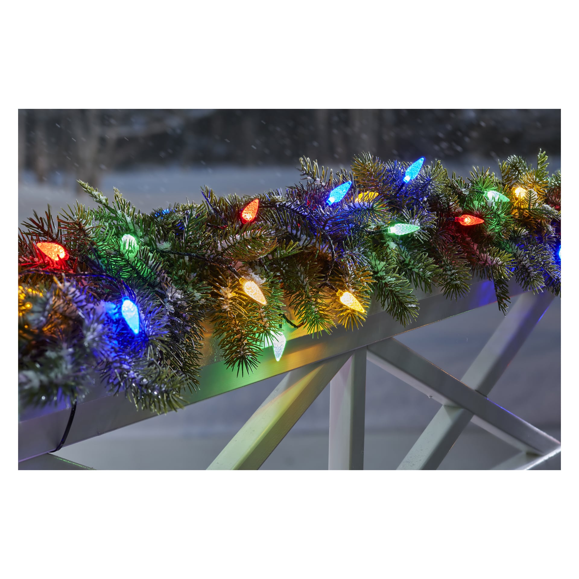EKVIP LED-ljusslingor med blå, röda, gula och gröna lampor är lindade runt en grön tallgirland som hänger över ett vitt räcke, med snötäckt landskap i bakgrunden.