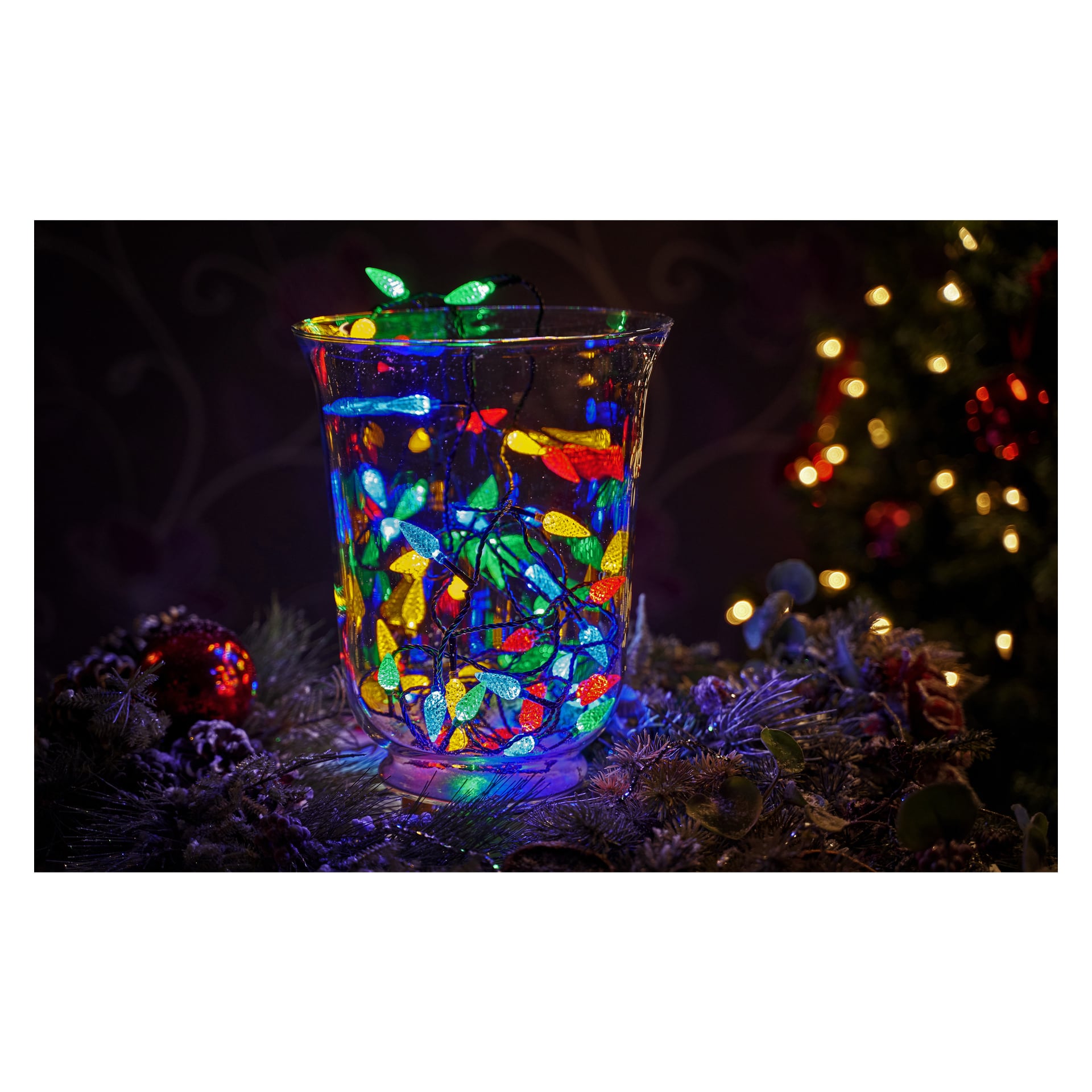 En glasvas med EKVIP LED-ljusslingor lyser på en festlig yta dekorerad med tallkvistar och prydnader, med en del av en tänd julgran synlig i bakgrunden.
