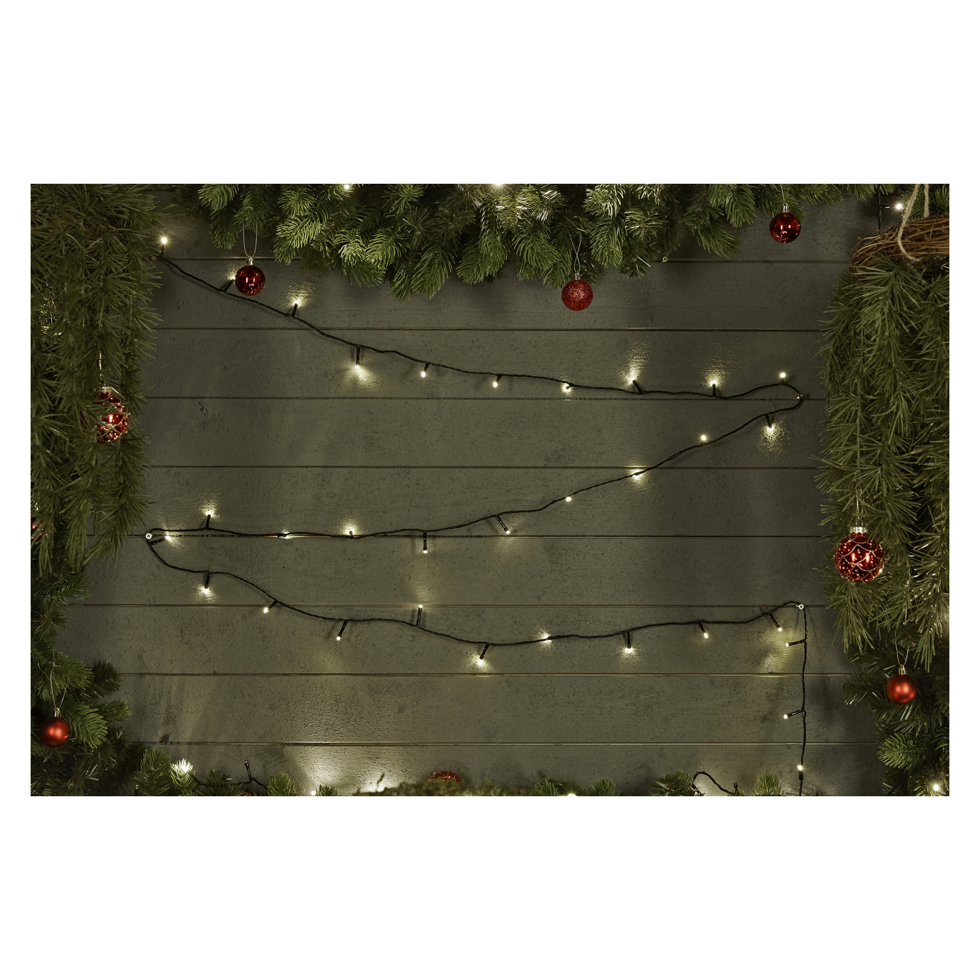 Batteriebetriebene EKVIP LED-Lichterketten und rote Ornamente hängen an einer dunklen Holzwand, eingerahmt von dicken grünen Kieferngirlandern. Die leuchtenden EKVIP-Lichter strahlen eine gemütliche Wärme aus und verstärken die festliche Dekoration, die die einladende Atmosphäre umgibt.