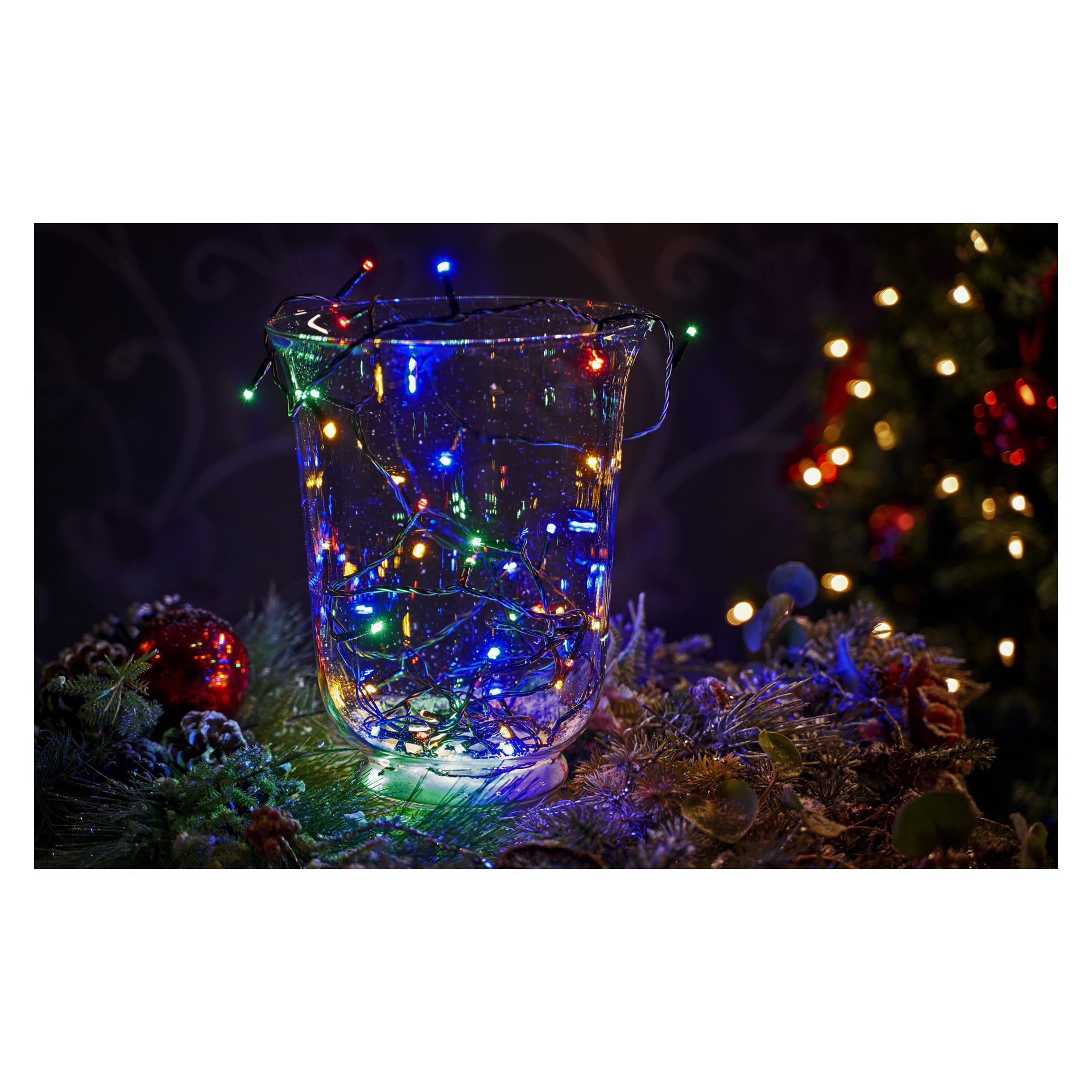 En klar glasvas fylld med en EKVIP batteridriven LED-ljusslinga placeras på en festligt dekorerad yta med tallgrenar och prydnader, med en lysande julgran synlig i bakgrunden.