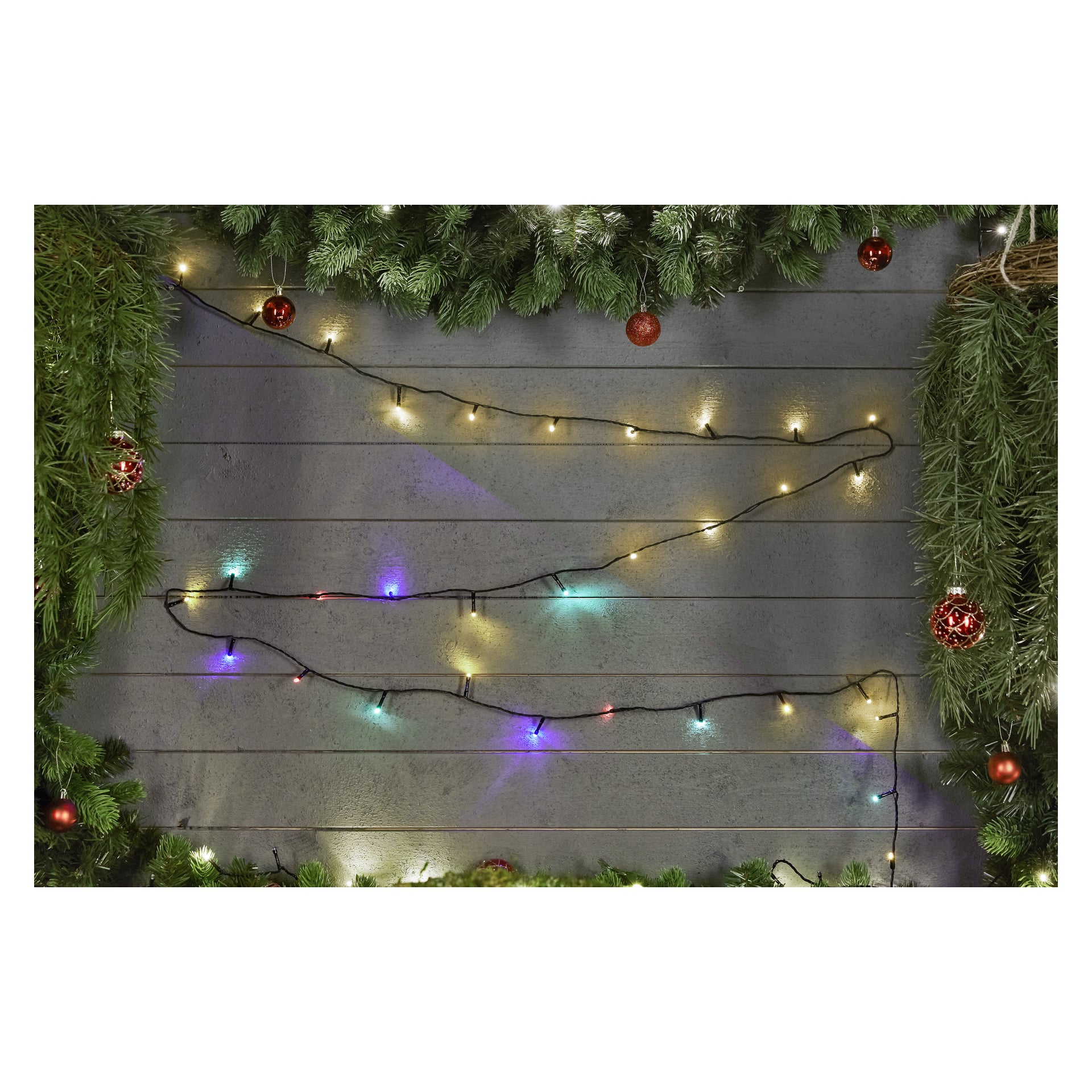 Gröna tallgirlanger och röda ornament pryder en trävägg, med två horisontella trådar av EKVIP-batteridrivna LED-ljusslingor – en med varmvita lampor och den andra med flerfärgade lampor – draperade över mitten.