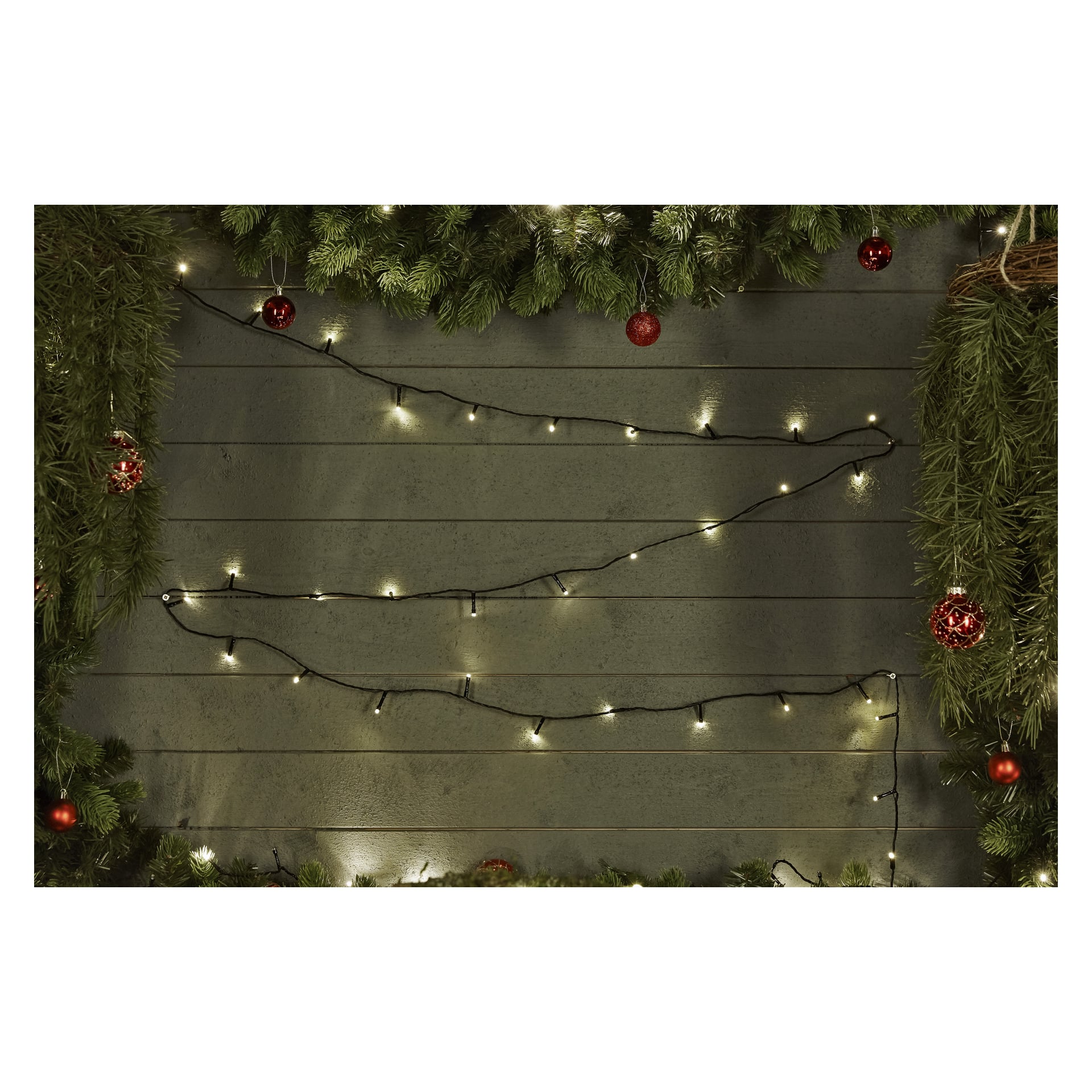 Batteridrivna EKVIP LED-ljusslingor och röda ornament pryder tallgirlanger som hänger runt en trävägg. EKVIP-lamporna lyser varmt, och träplankorna i mitten av scenen löper horisontellt.
