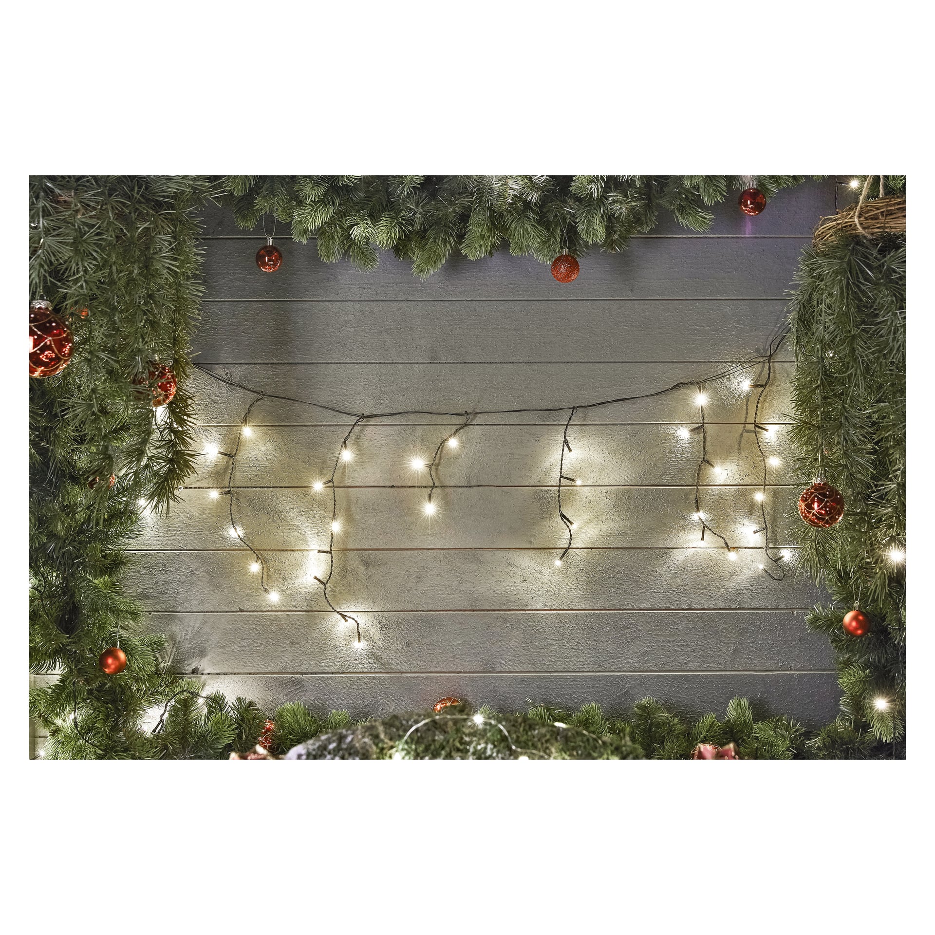 En trävägg kantad med grangrenar är dekorerad med EKVIP:s LED-istappssträng Connectable System och röda ornament. De upplysta ljusen bidrar till en festlig högtidsstämning.