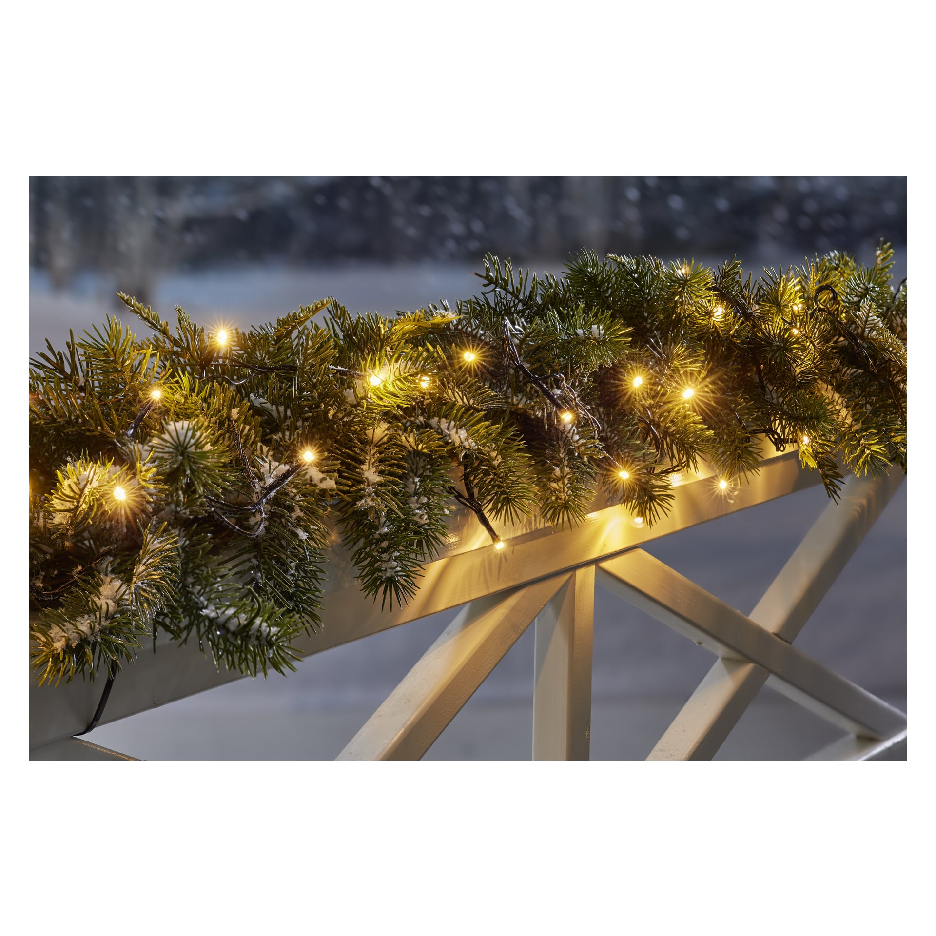 En rad EKVIP LED-lampor är vävd genom snötäckt grön girland och placerad över ett vitt träräcke, med ett mjukt suddat vinterlandskap bakom.