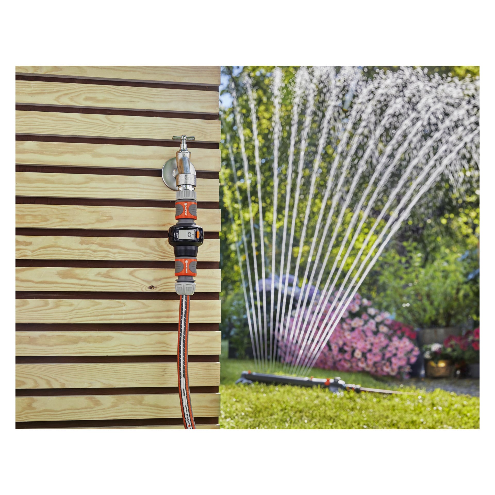 En Gardena AquaCount vattenmätare med en orange och grå kontakt är kopplad till en trädgårdskran monterad på en trävägg. Bakom den sprutar vatten från en sprinkler över gräset och blomsterbäddarna.