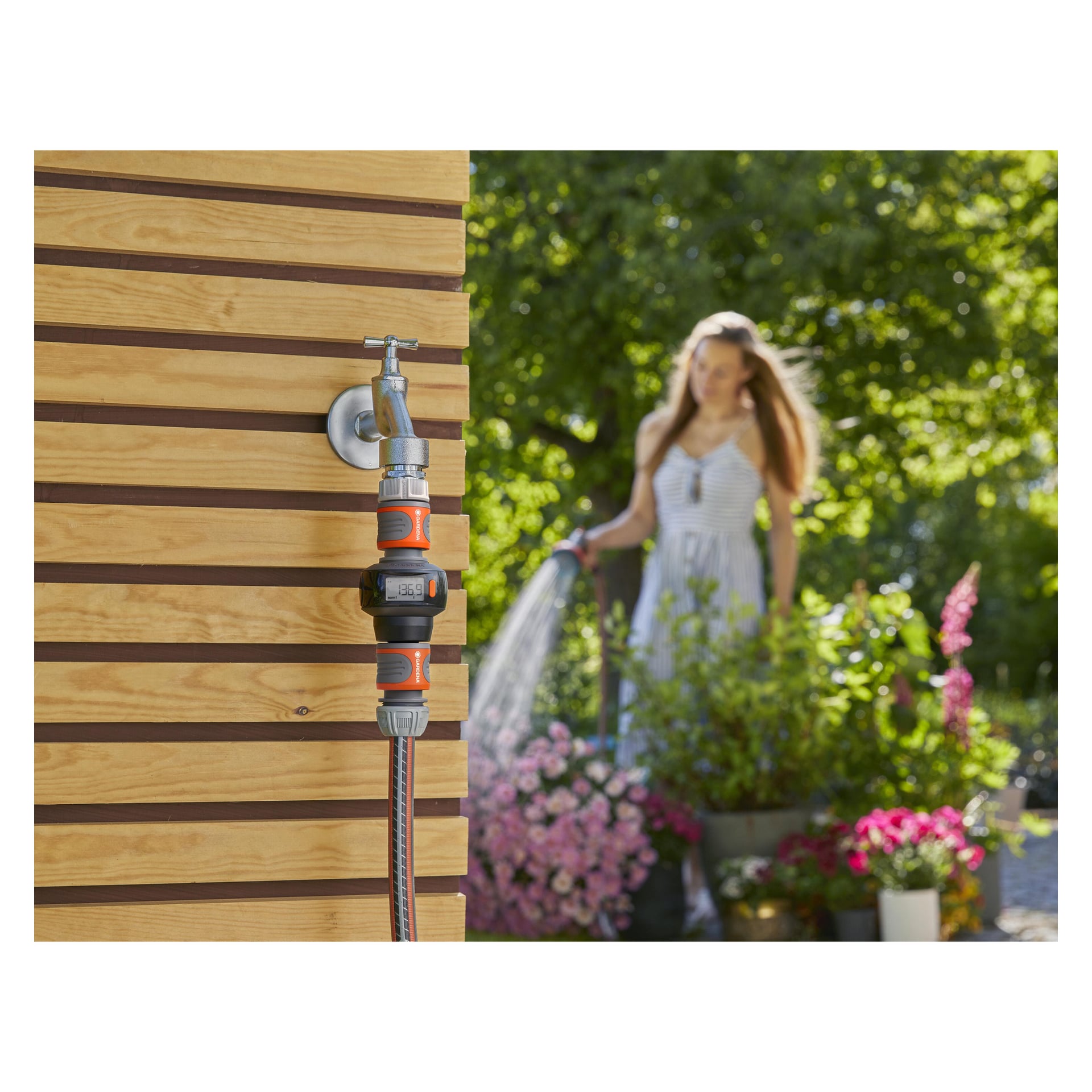 En Gardena Water Meter AquaCount är fäst vid en utomhuskran på väggen. I den mjukt suddiga bakgrunden vattnar en kvinna i randig klänning växter och blommor omgiven av frodig grönska.