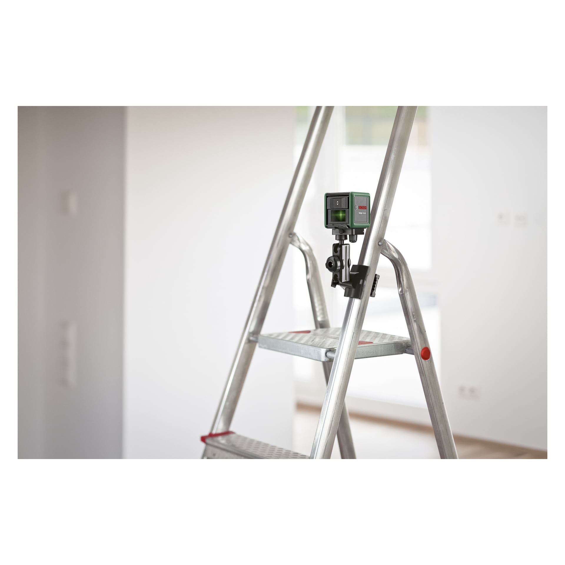 Et Bosch Quigo Green laservater er festet til en metallstige i et godt opplyst, tomt rom med hvite vegger og store vinduer.
