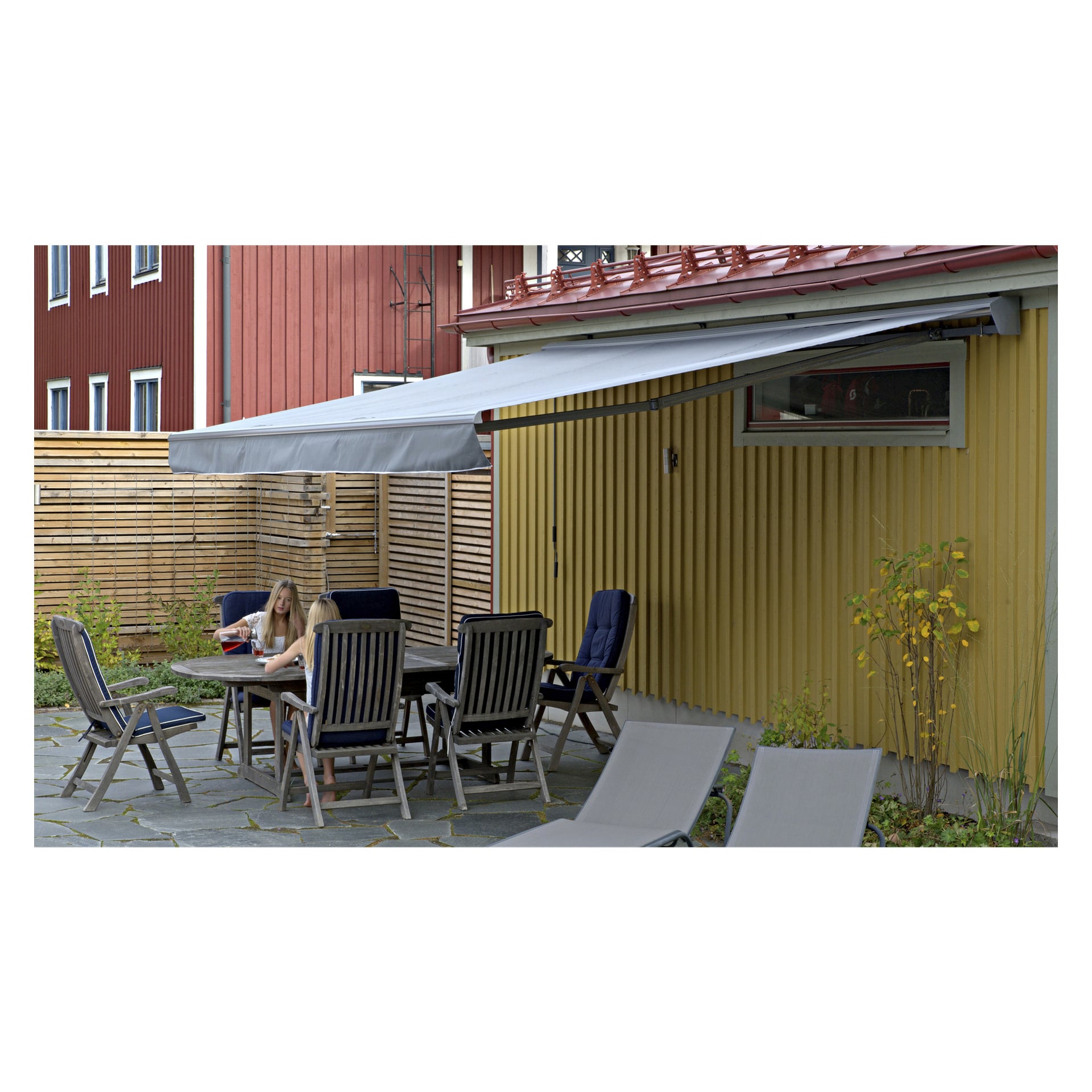 Två personer sitter vid ett utebordsbord med stolar under en grå Hard Head Patio-markis fäst vid ett gult hus. Uteplatsen har stengolv, flera solstolar och är omgiven av ett trästaket.