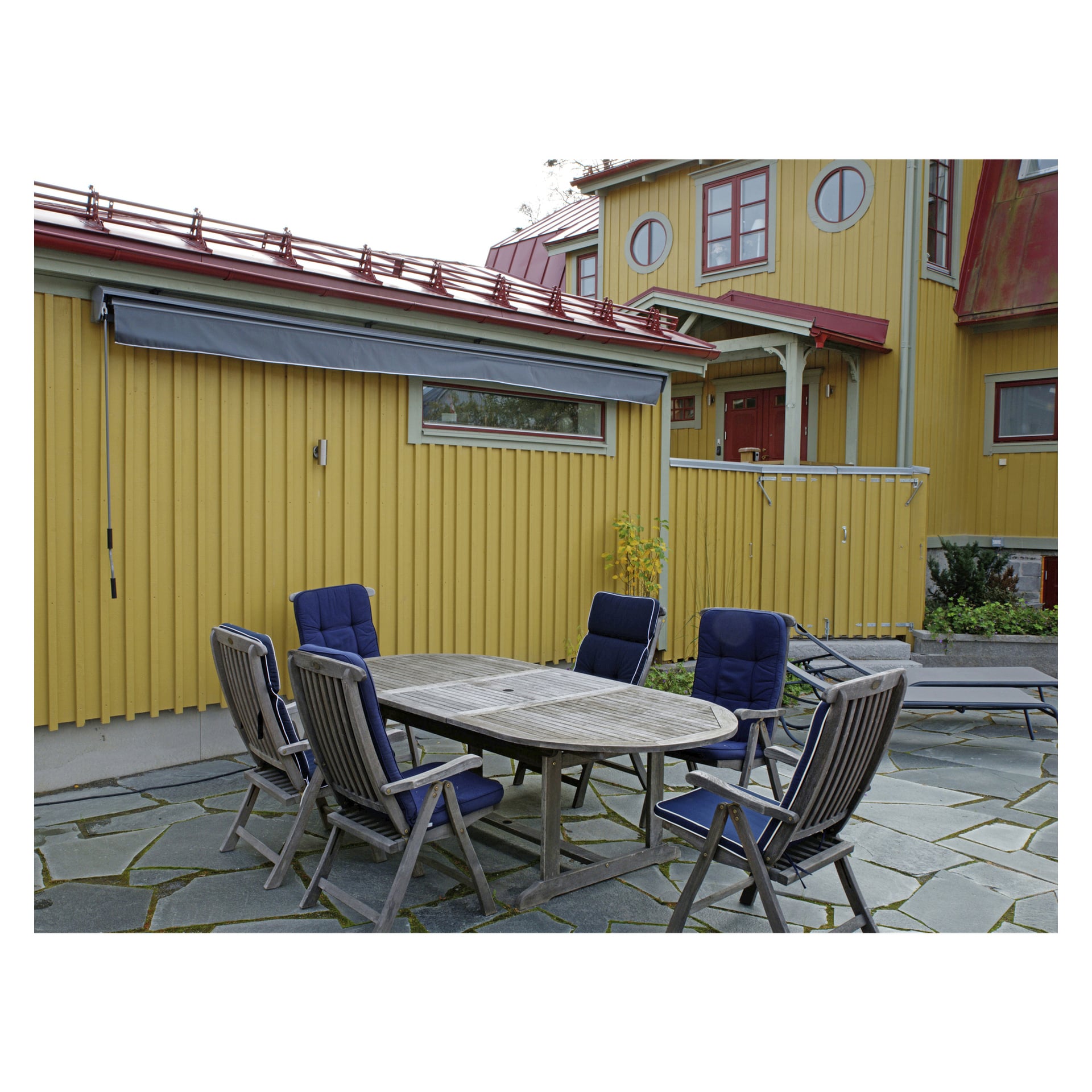 Ett ovalt träbord med sex blådämpade stolar är placerat på en stenplatta framför ett gult hus med röda lister. Ovanför uteplatsen skuggar en Hard Head Patio-markis fönstret.
