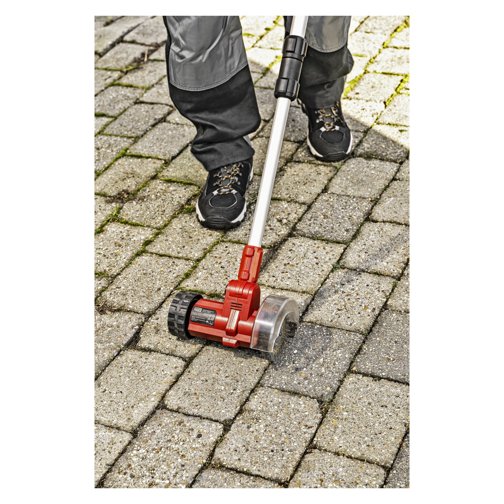 Man bedient den Meec Tools Multiseries Paving Scraper PW-18V HOME 300, um Unkraut aus den Lücken zwischen den Pflastersteinen auf einem Außenweg zu entfernen, wobei nur die Unterschenkel und schwarzen Schuhe sichtbar sind.