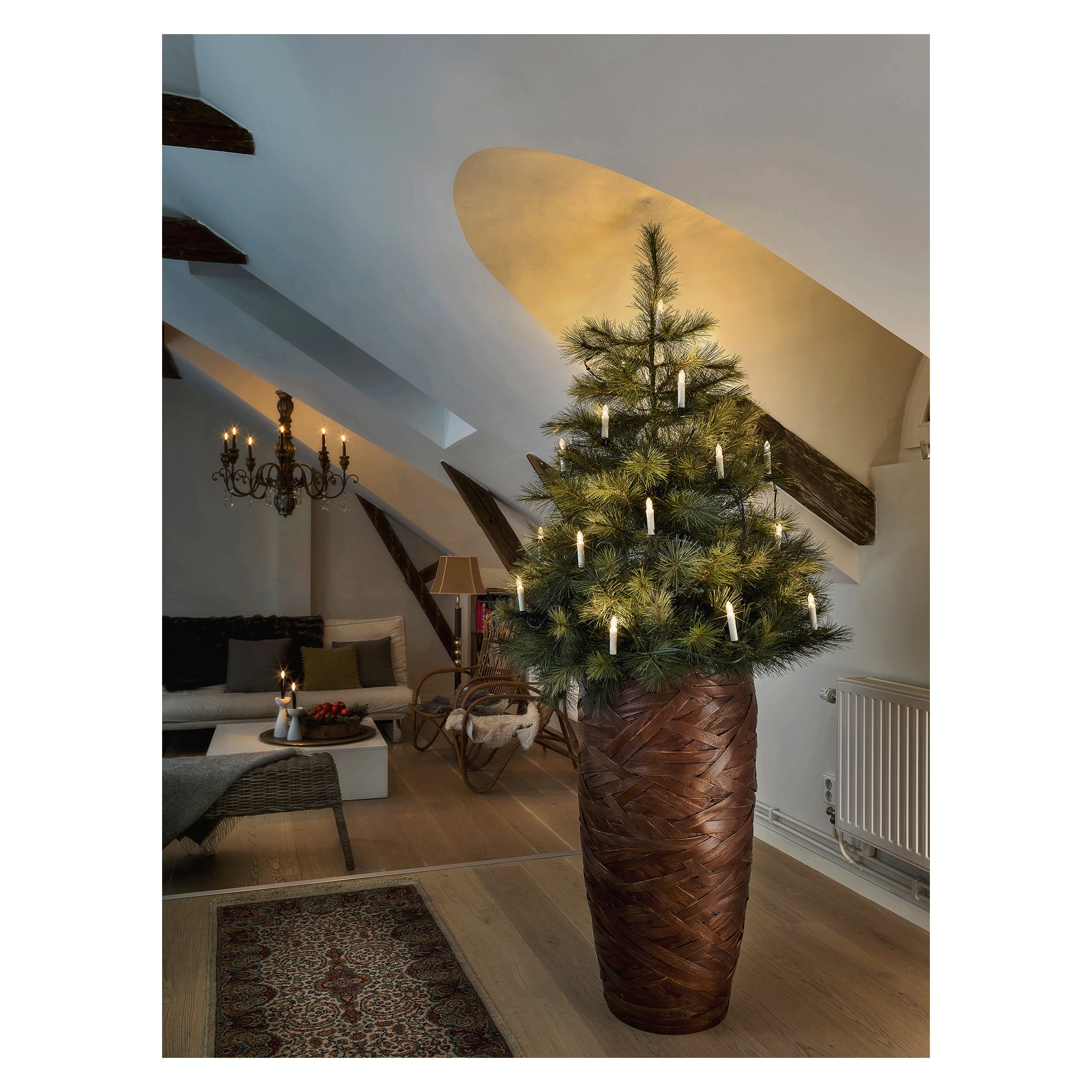 En hög konstgjord julgran prydd med EKVIP LED-lampor står i en stor vävd kruka i ett varmt vardagsrum med trägolv, synliga balkar, en ljuskrona, en soffa, en matta och en synlig radiator.
