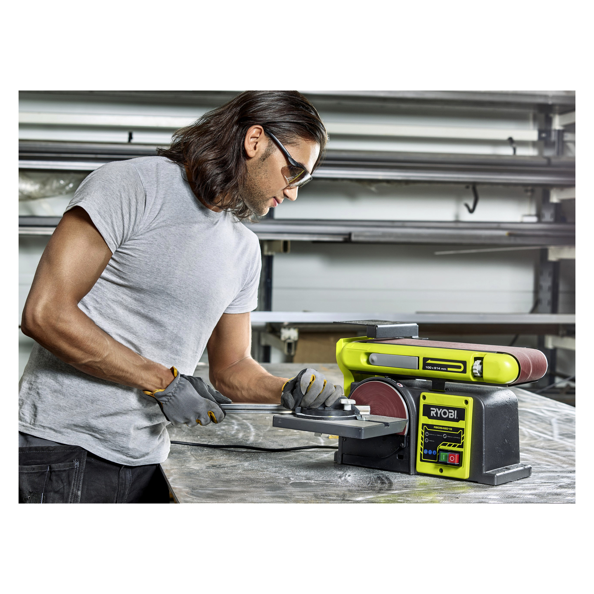 En person med vernebriller og hansker sliper en metallbit på et bord ved hjelp av Ryobi-beltet/skivesliperen RBDS4601G i et verksted, med ulike verktøy og metallrør synlige rundt seg.