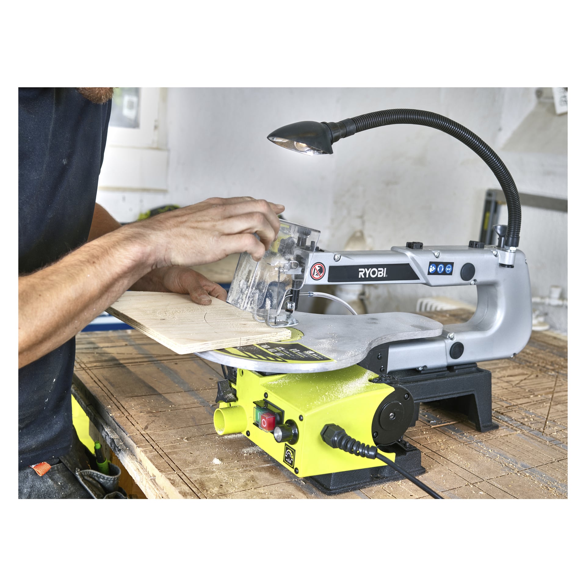 Någon använder en Ryobi-scrollsåg RSW1240G för att såga en lätt träbit i en verkstad. Den gröna och grå Ryobi-märkta sågen vilar på bordet, med trädamm utspritt i närheten. Operatörens ansikte är utom synhåll.