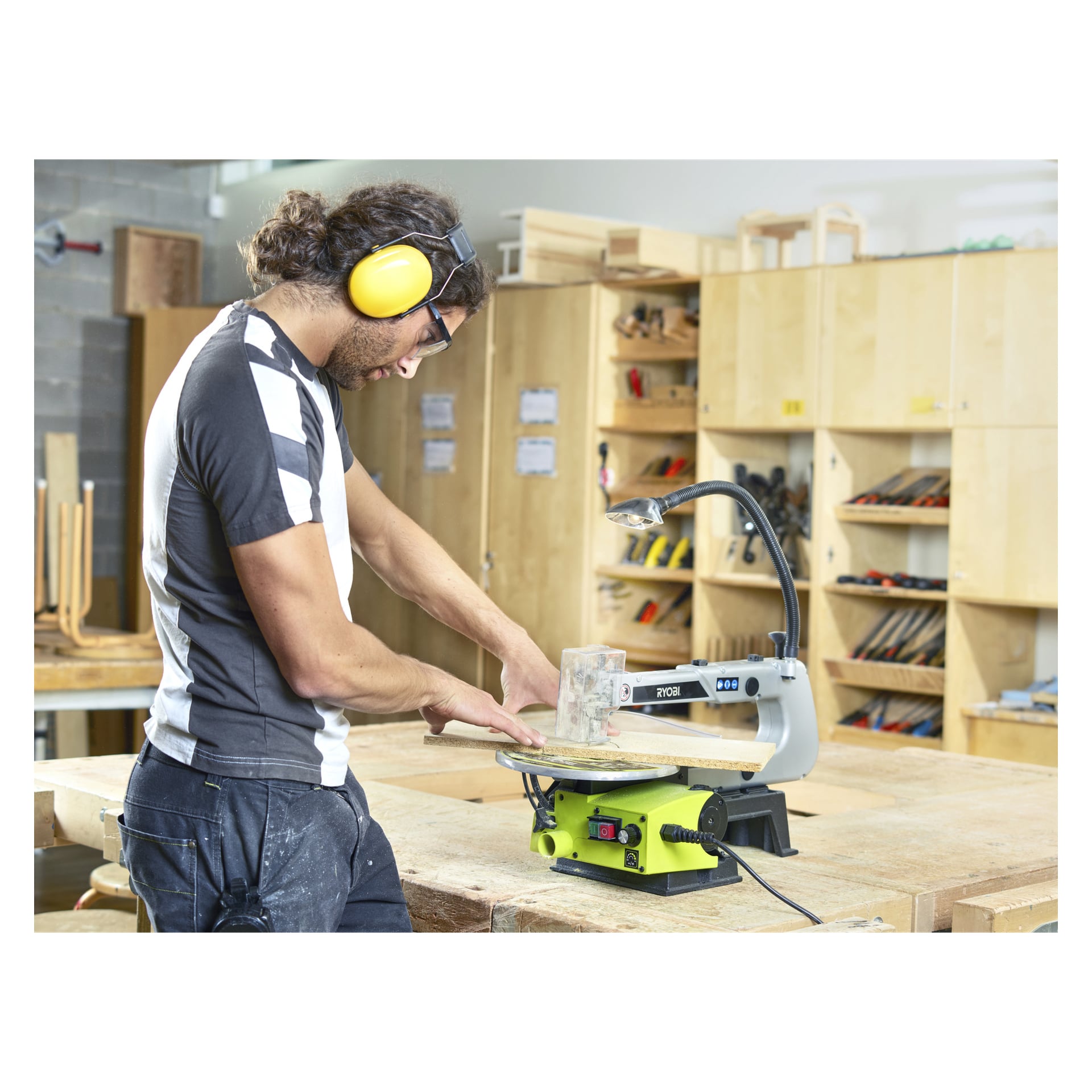 Med skyddsöronskydd använder man Ryobi-scrollsågen RSW1240G för att såga trä i en verkstad. Hyllor med verktyg och material kan ses bakom dem när de koncentrerar sig på sitt arbete.