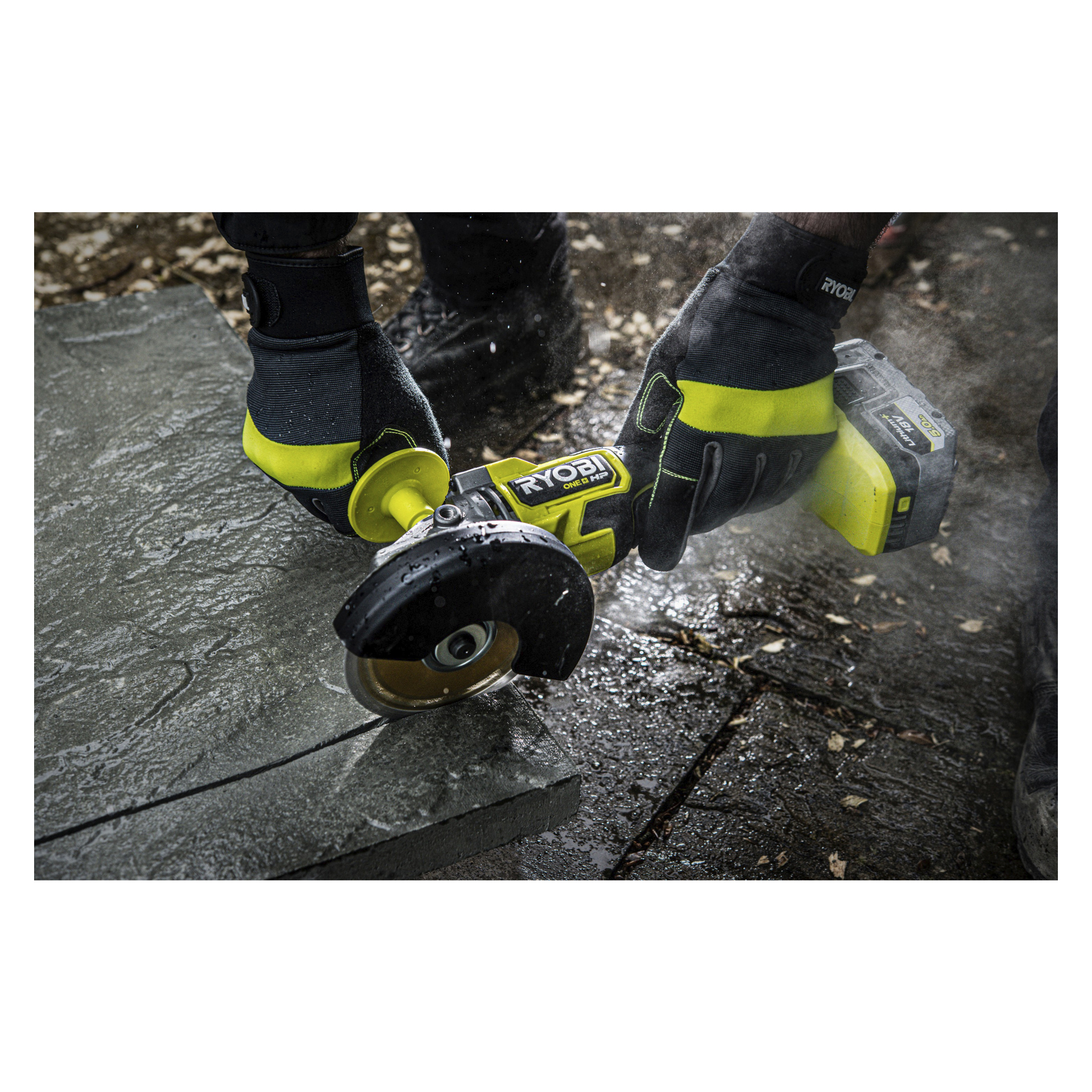 Osoba w czarno-żółtych rękawiczkach obsługuje szlifierkę kątową Ryobi One+ HP RAG18X-140SJ, aby wycinać betonową płytę, tworząc kurz z rozsypanymi liśćmi i płytkami na ziemi.