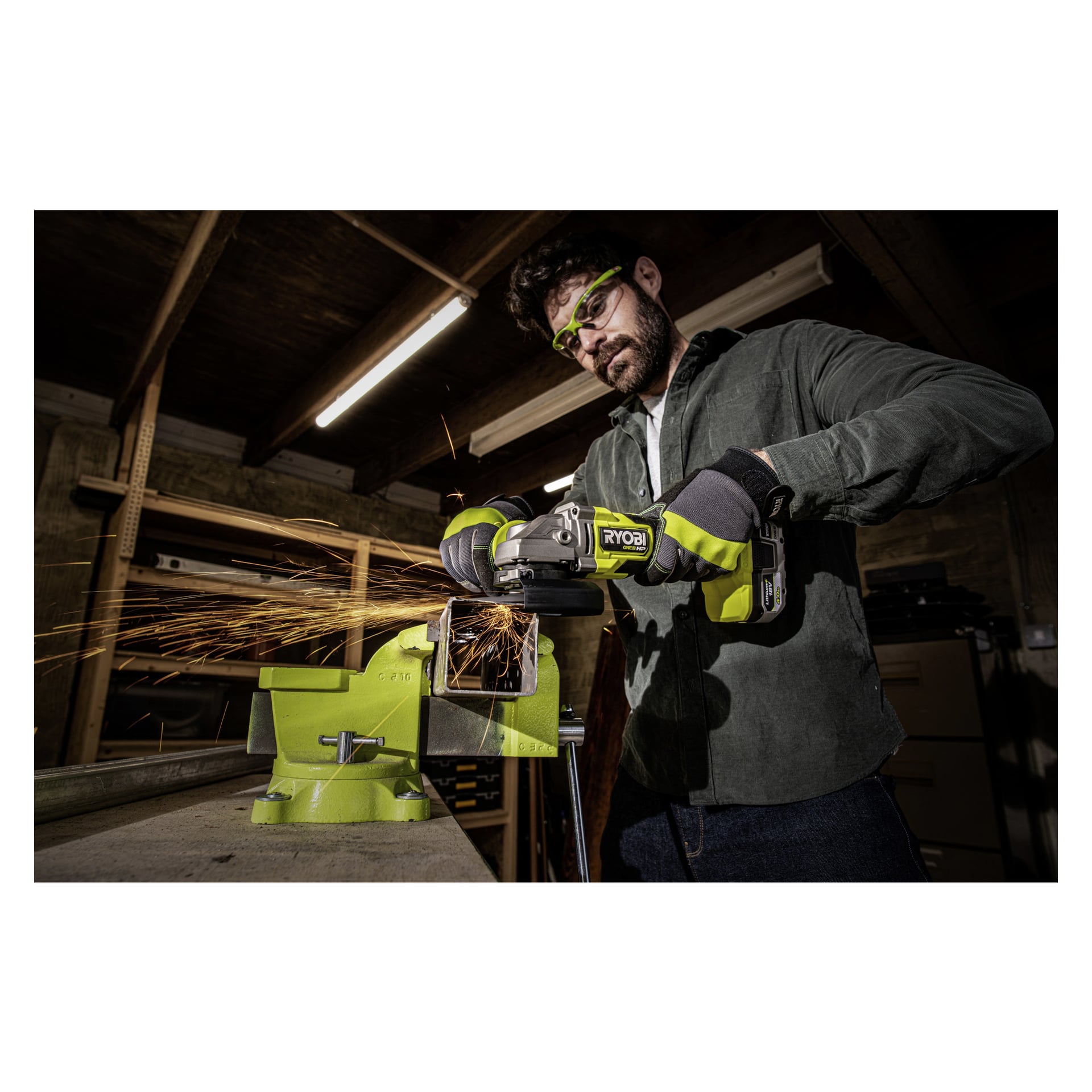 En person, skyddad av skyddsglasögon och handskar, använder en Ryobi One+ HP vinkelslipare RAG18X-140SJ för att skära en metallbit som är fastklämd i ett skruvstäd. Gnistor flyger runt i verkstaden, som har träbjälkar och hyllor i bakgrunden.