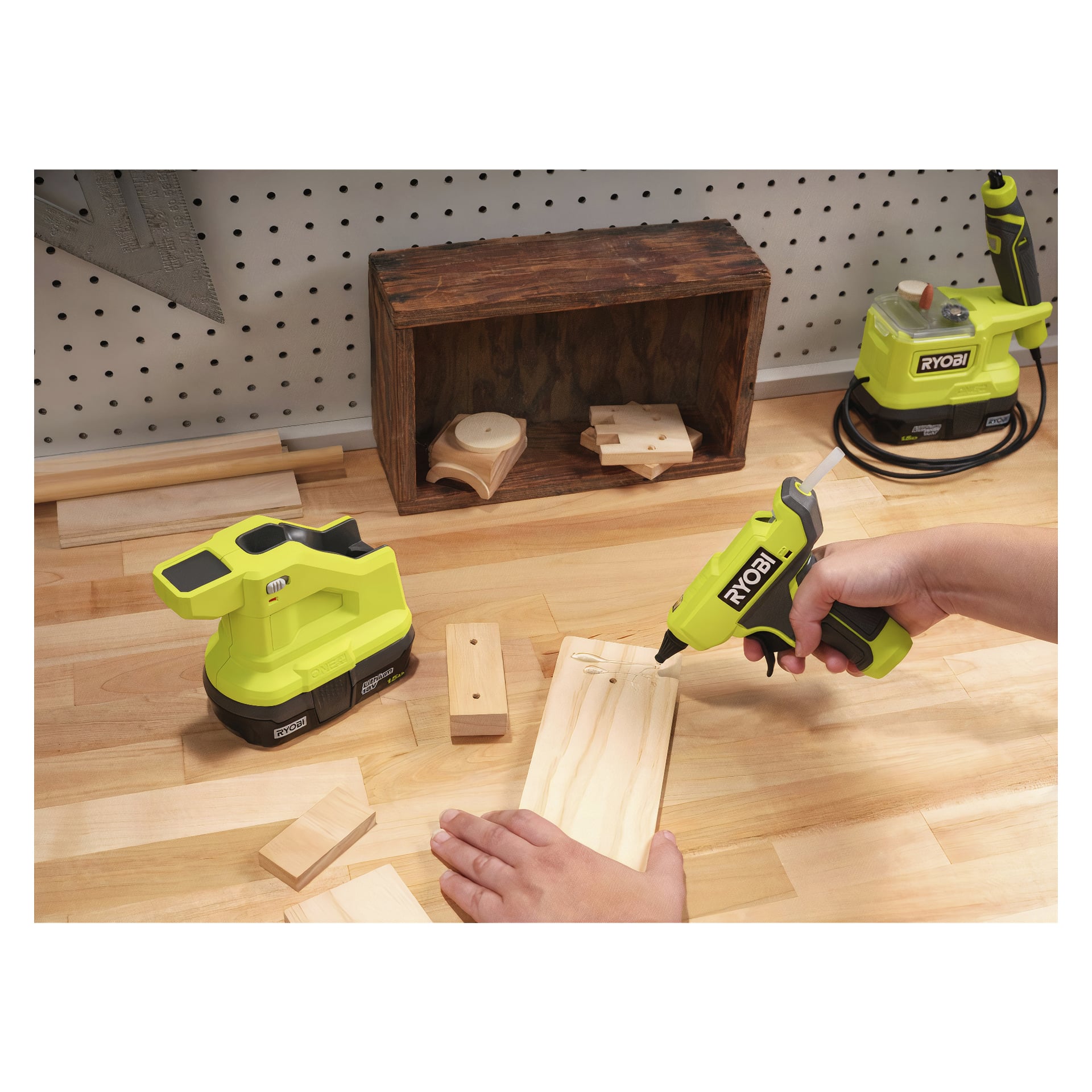 Någon applicerar lim på en träbit på en arbetsbänk med en Ryobi One+ batteridriven limpistol RGLM18-0. I bakgrunden kan man se fler träklossar och Ryobi One+-verktyg.