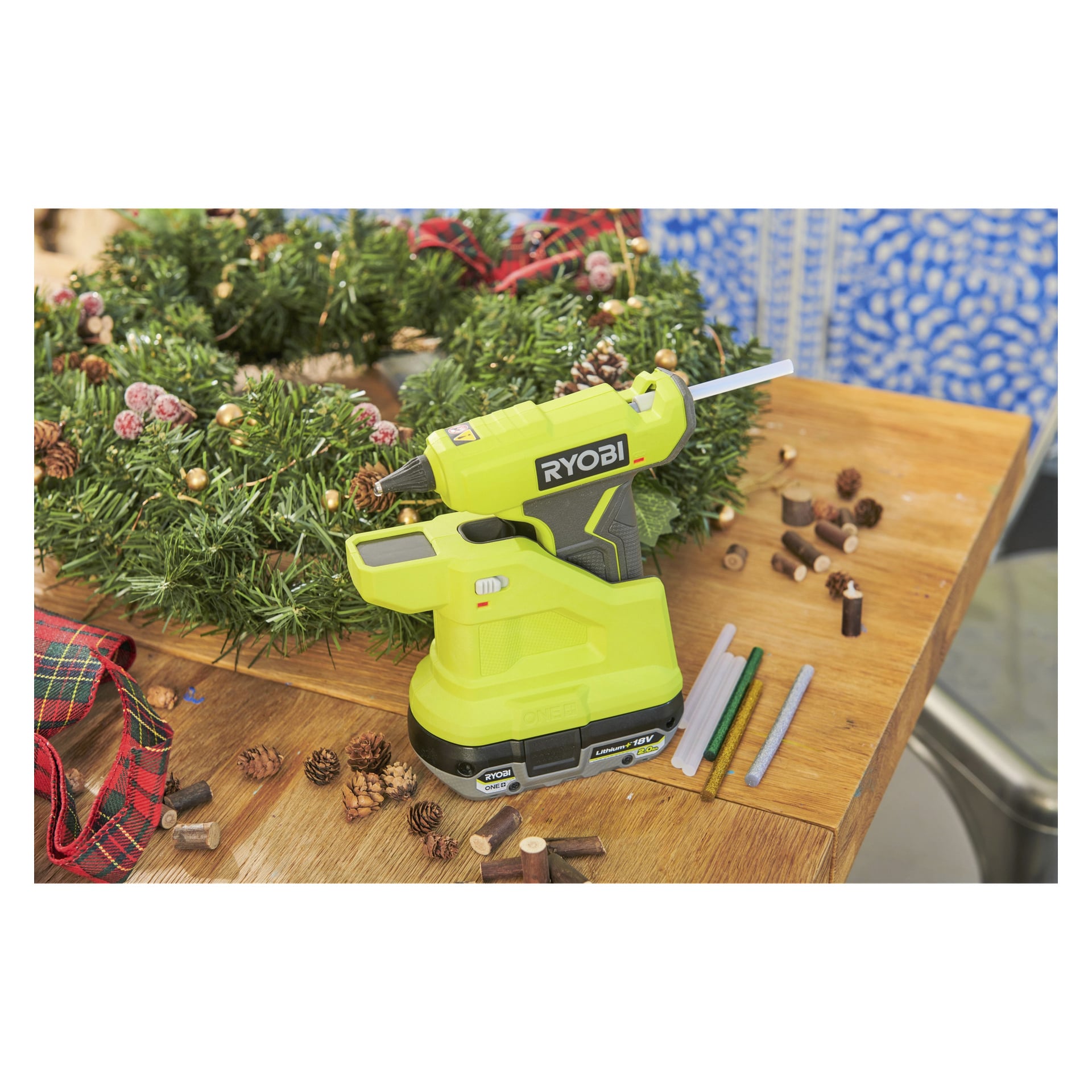 En gul Ryobi One+ batteridriven limpistol RGLM18-0 vilar på ett träbord omgivet av limstift, en festlig julkrans, kottar, band och diverse pysselmaterial.