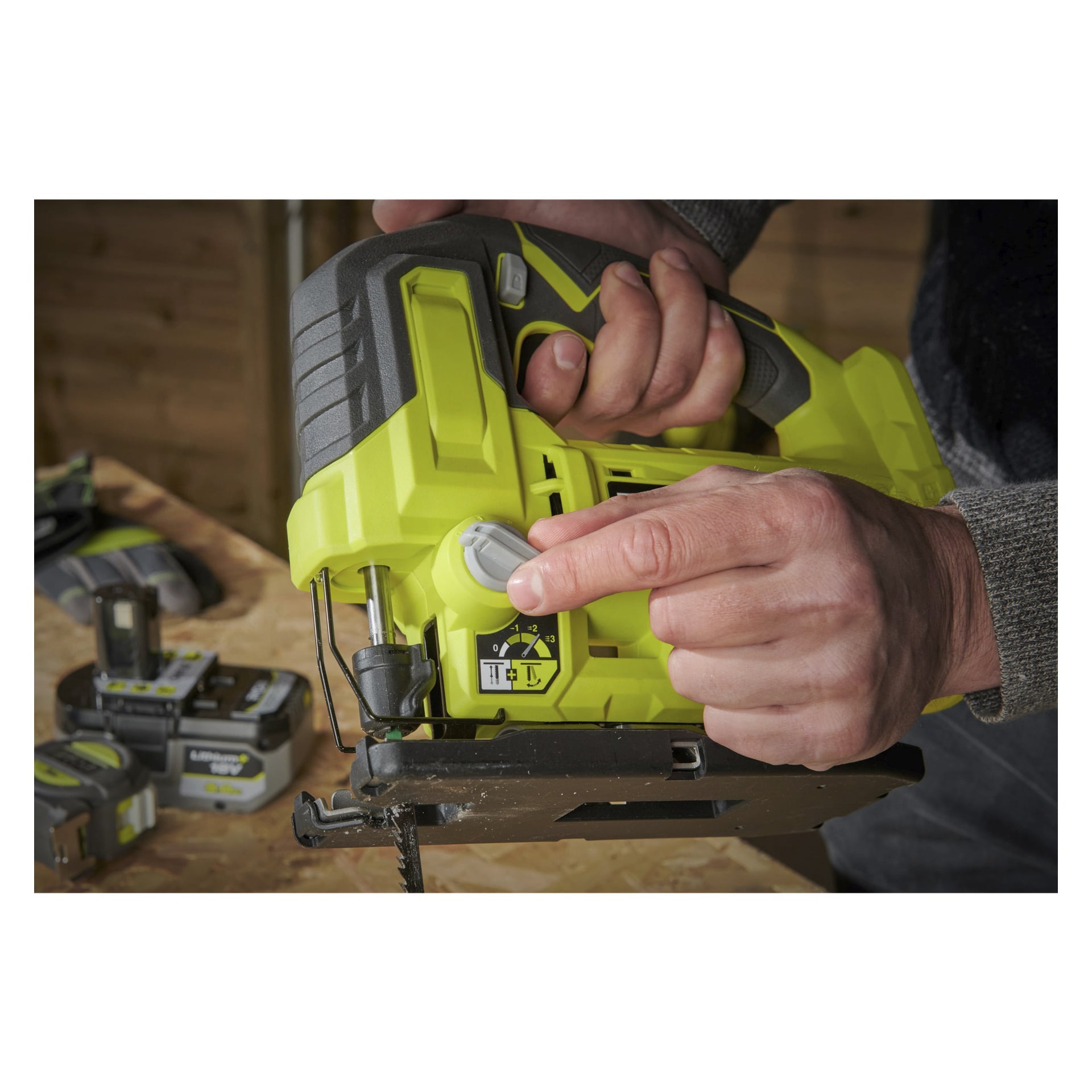 Någon justerar kontrollerna på en batteridriven sticksåg RJS18-0 av Ryobi One+, placerad ovanför en träbit. Flera andra Ryobi One+-verktyg och batteripaket kan ses bakom dem.