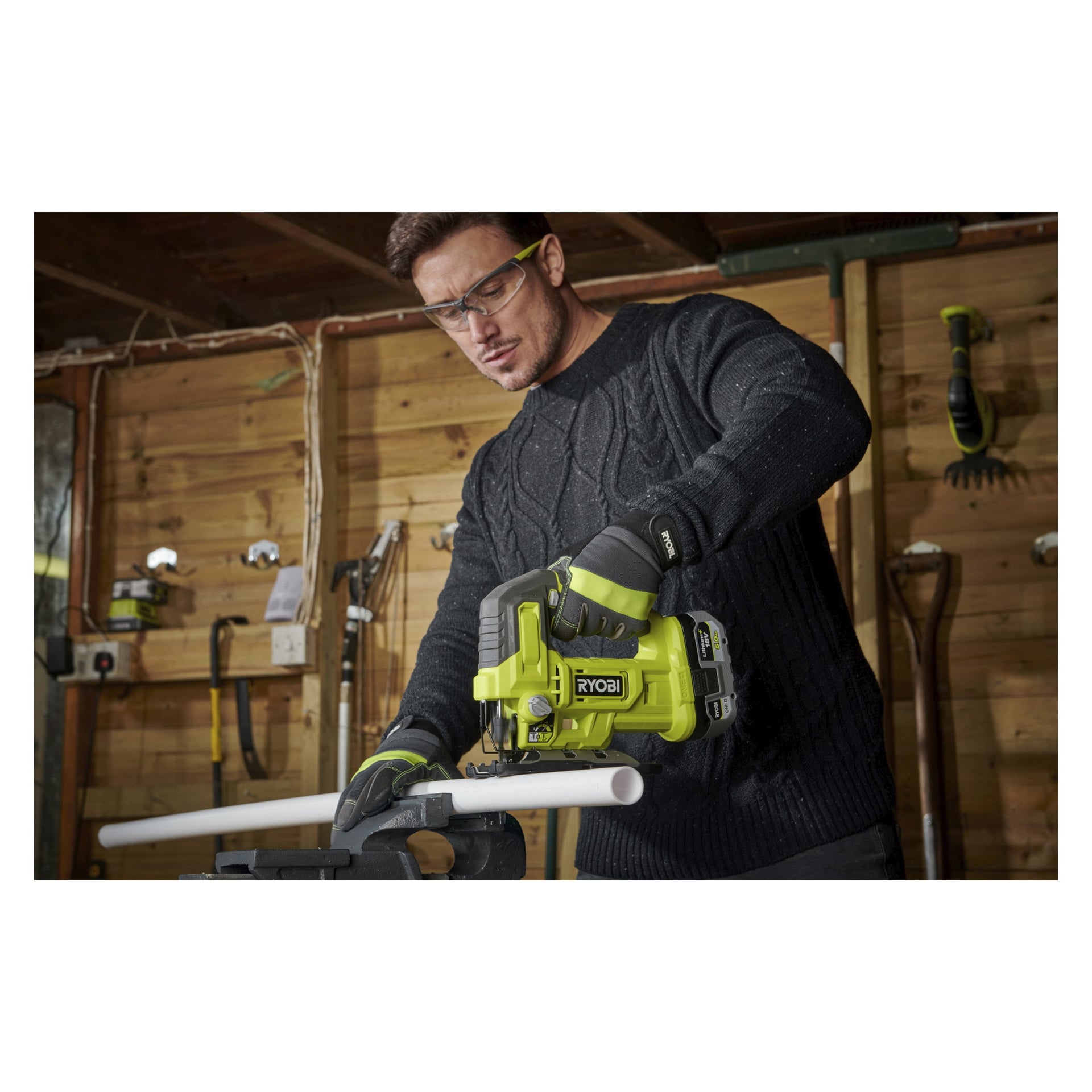 Med skyddsglasögon och handskar använder en person en grön Ryobi One+ batteridriven sticksåg (modell RJS18-0) för att skära ett vitt PVC-rör på en arbetsbänk i en träverkstad, med olika verktyg hängande på väggen bakom dem.