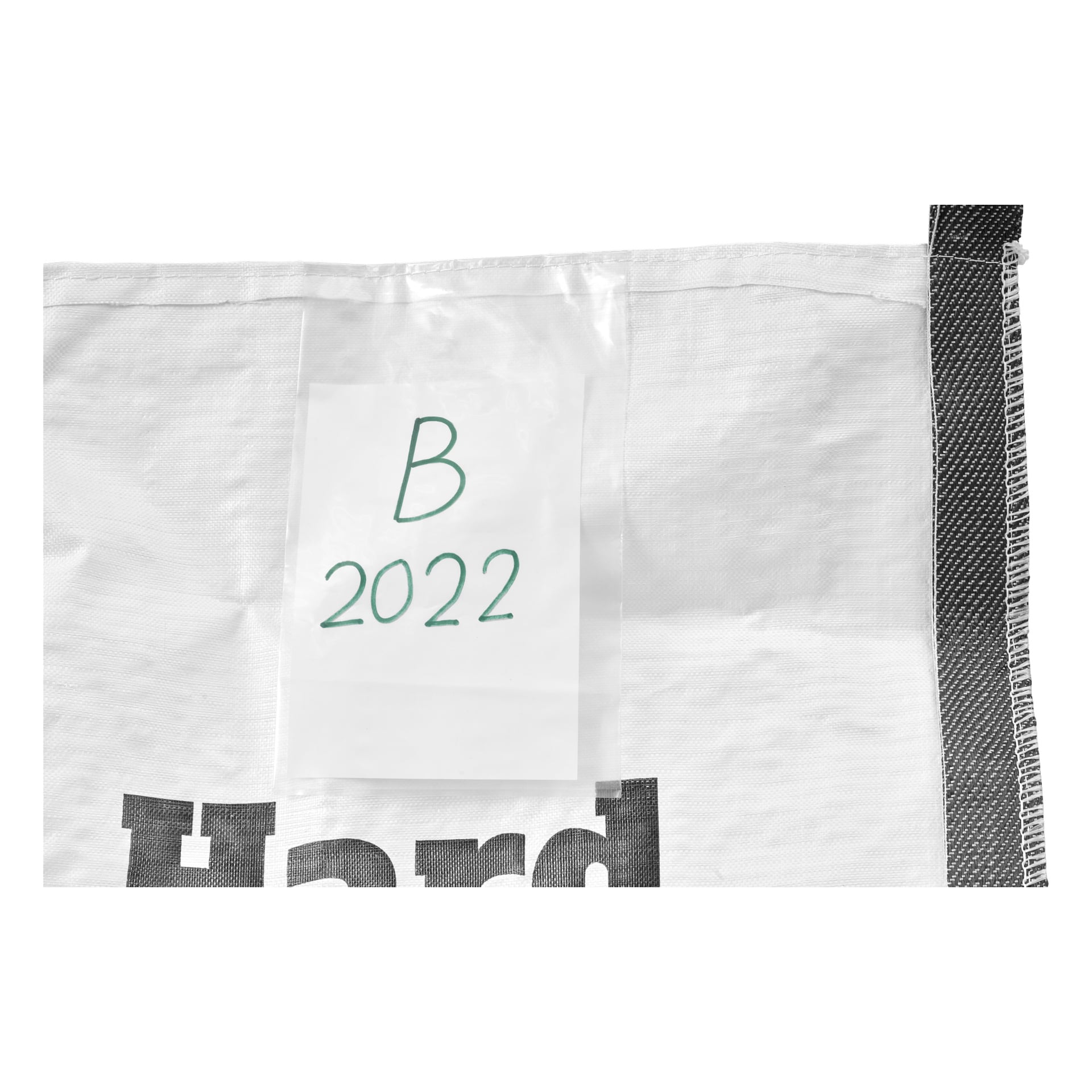 En Hard Head Builders-väska med vit tygyta och svart kantremsa visas. En transparent ficka innehåller ett papper märkt "B 2022" med grön handstil, och delvis svarta tryckta bokstäver visas längst ner på bilden.