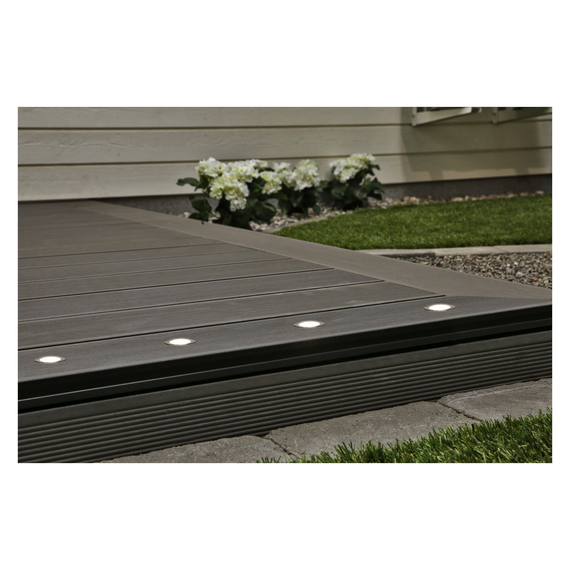 En närbild av en grå kompositaltan visar fyra Anslut Decklights LED-lampor från en 10-pack installerad längs kanten, med grönt gräs, vita blommor och dekorativa stenar som kantar däcket nära en beige fasadvägg.