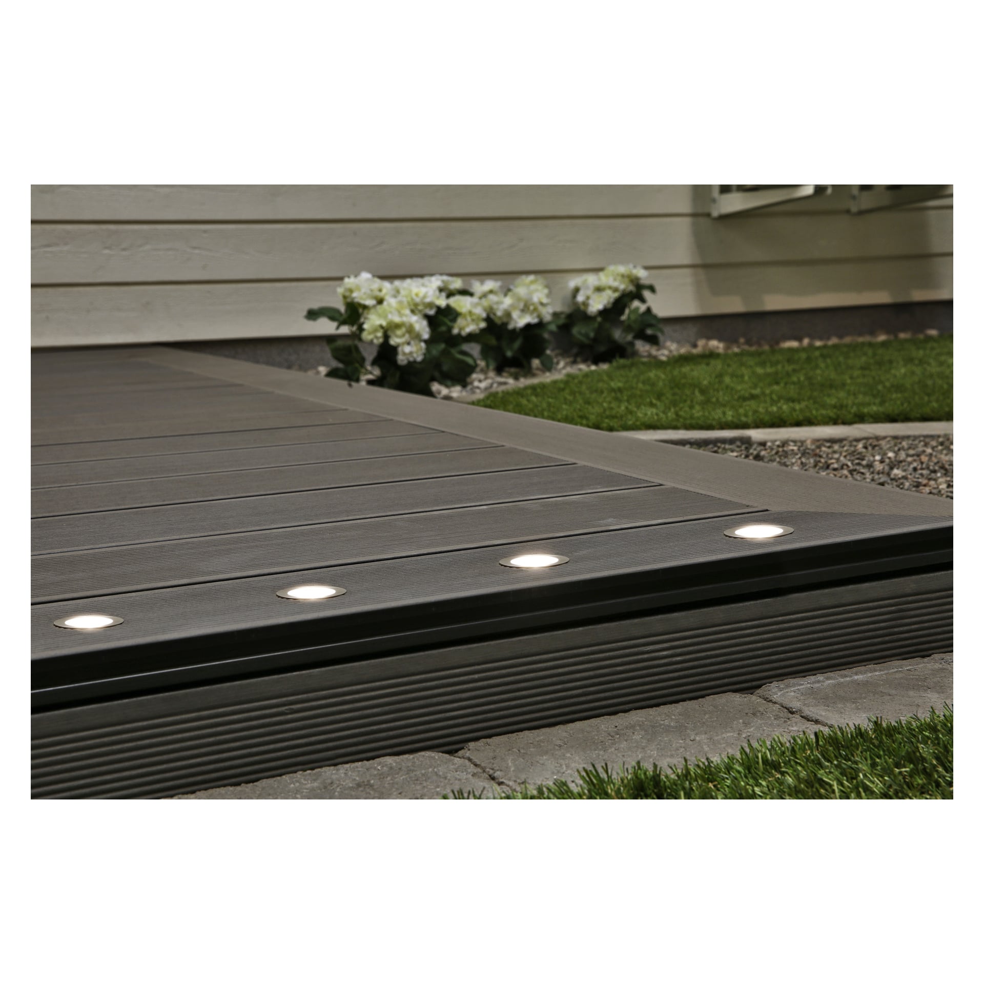 En altankant upplyst av integrerade runda LED-lampor från Anslut Decklights LED 10-pack, omgiven av gräs, vita blommor och små dekorativa stenar bredvid en beige husvägg.