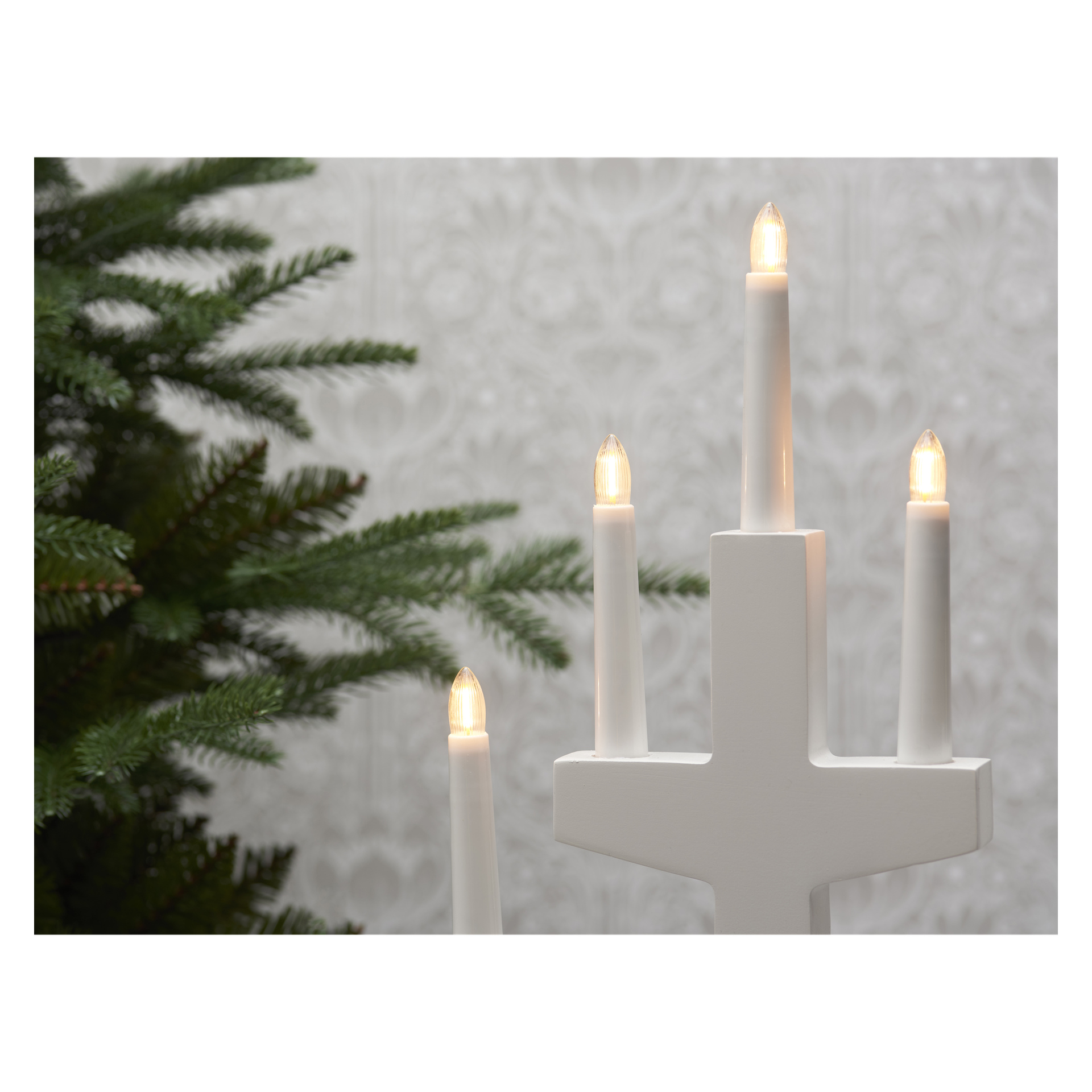 En EKVIP vit adventsljusstake med fyra lysande LED-ljus och reservlampor från EKVIP Spare Bulbs for Advent Candlestick LED 3-pack står bredvid gröna tallgrenar. En subtilt mönstrad vägg syns i bakgrunden.