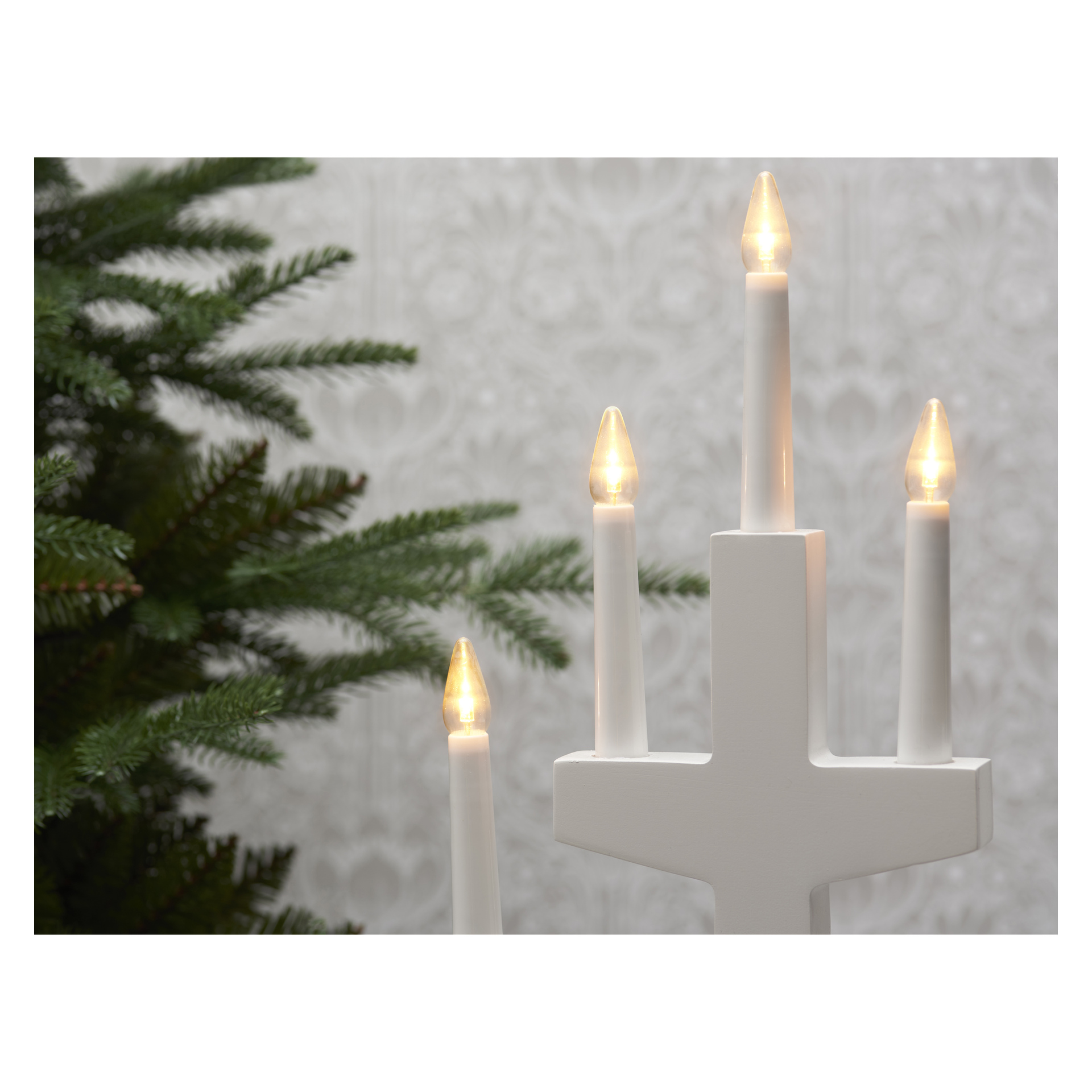 En upplyst vit adventsljusstake med EKVIP LED-ljus visar fem lysande lampor bredvid gröna tallgrenar, placerade framför en mönstrad vägg. Trepack reservlampor finns tillgängliga för denna ljusstake.
