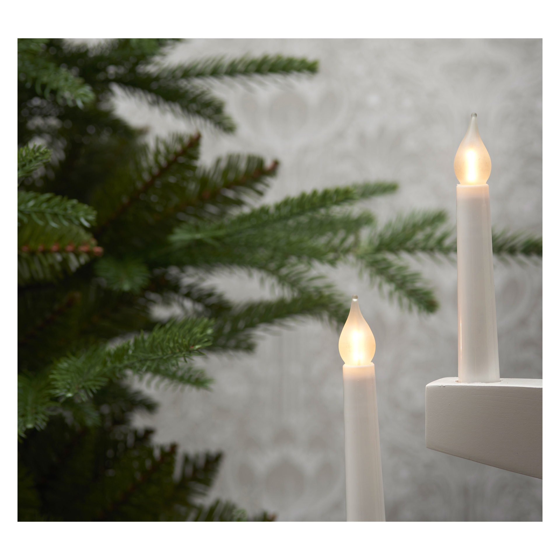 Tre ersättningslampor för en EKVIP Advent-ljusstake är placerade framför en grön konstgjord tall, med en subtil mönstrad bakgrund bakom dem.