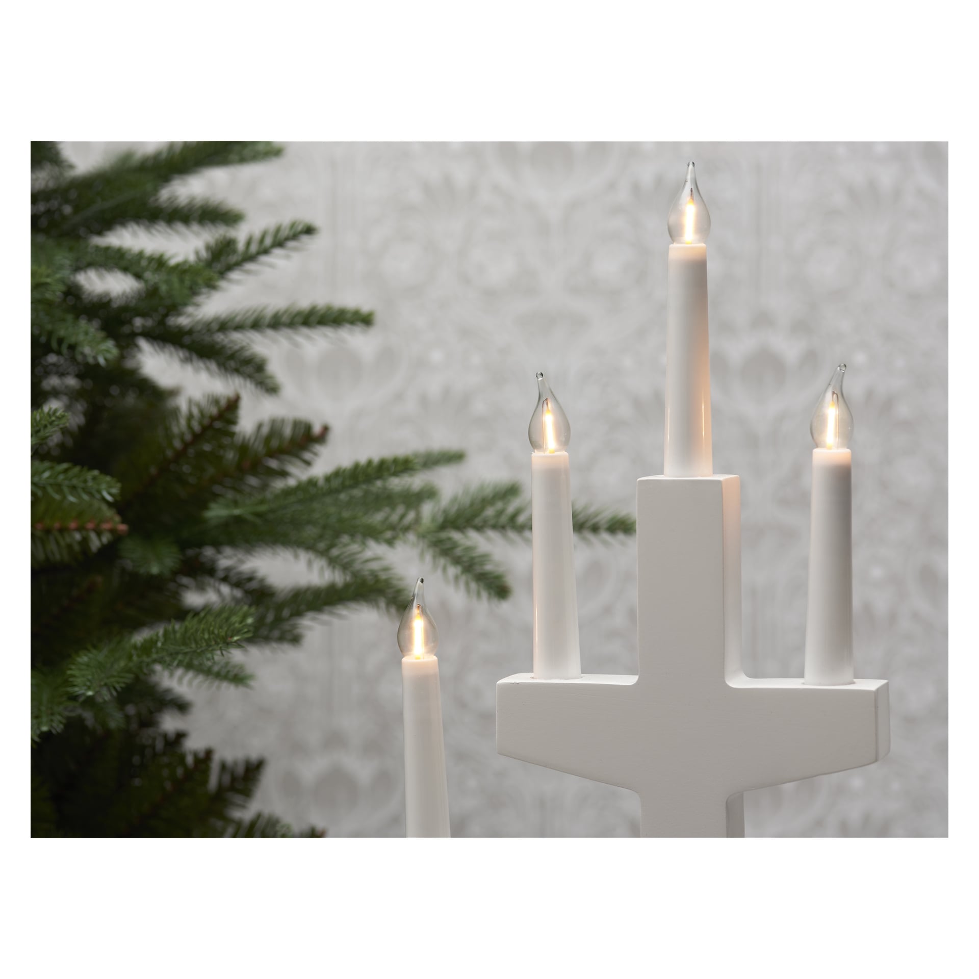 En EKVIP Advent-ljusstake med fyra lysande LED-ljus och kompatibla reservlampor (3-pack) står framför en mönstrad vägg, med gröna tallgrenar synliga till vänster.