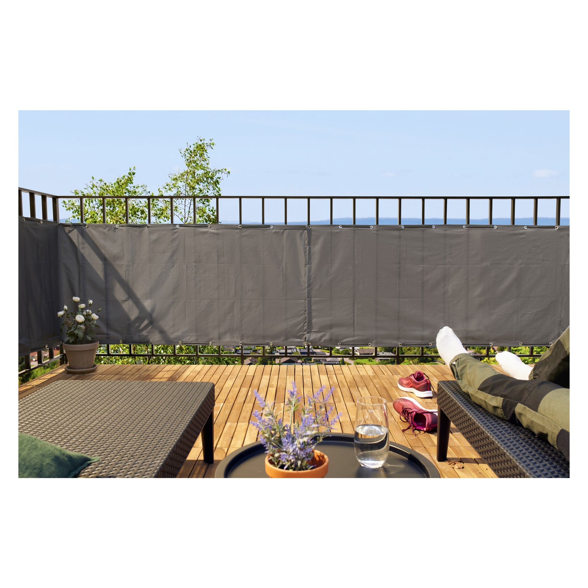 Någon slappar bekvämt på en stol på en träbalkong med Hard Head Balcony insynsskärm och frodiga krukväxter. Bredvid dem står röda skor och ett glas vatten, med utsikt över öppen himmel och avlägset vatten i bakgrunden.