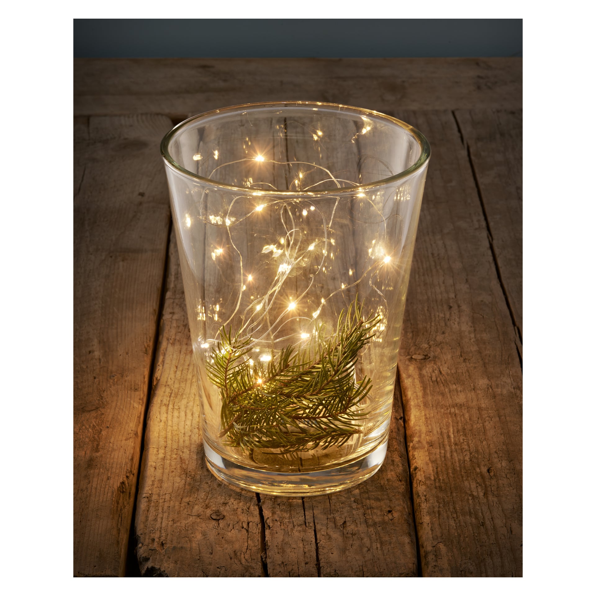 En klar glasvas med en EKVIP-batteridriven LED-ljusslinga och en liten tallgren vilar på en rustik träyta och utstrålar ett varmt, milt sken.