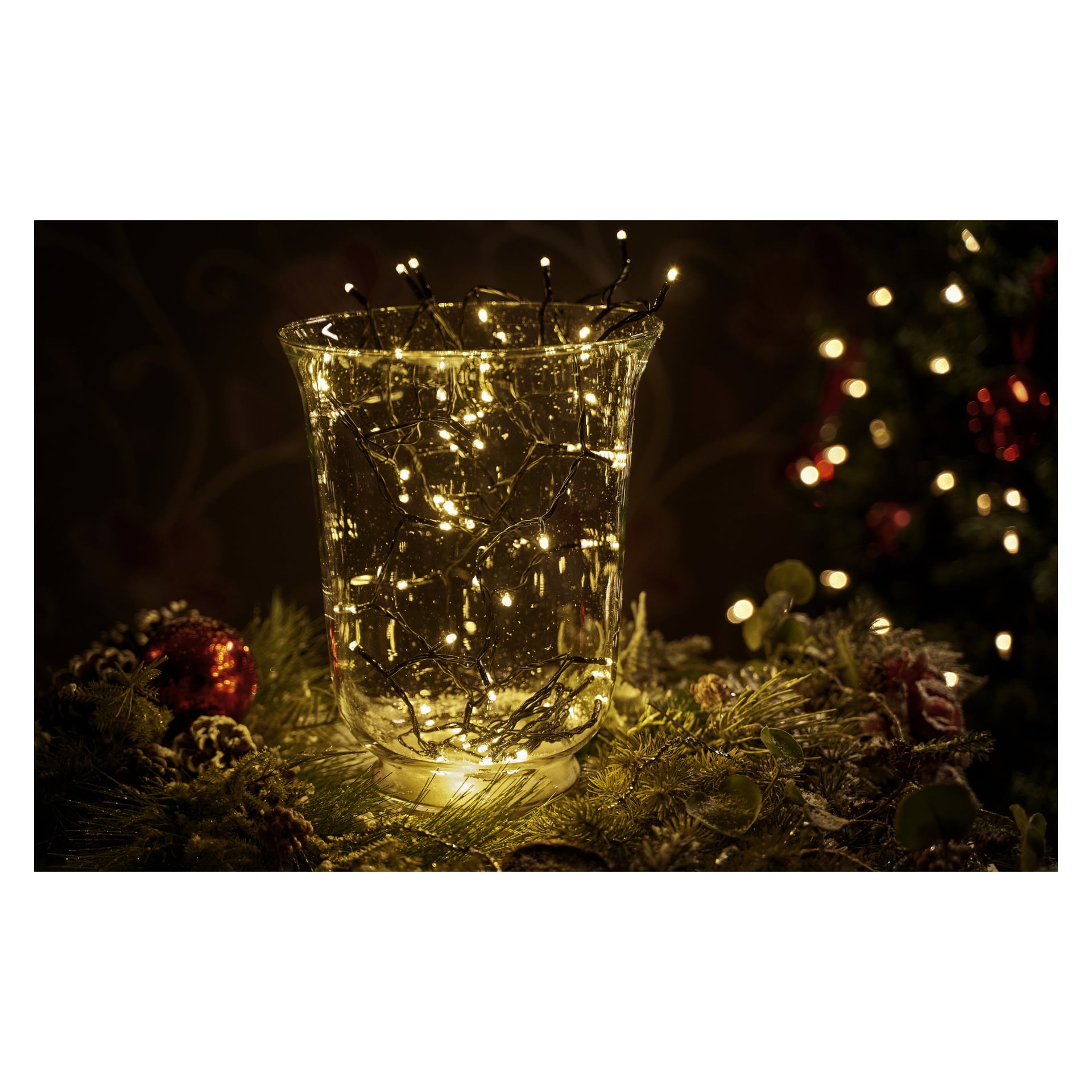 Eine klare Glasvase, gefüllt mit EKVIP LED-Lichterketten, steht auf einer Oberfläche, die mit Kiefernzweigen, Kiefernzapfen und roten Ornamenten geschmückt ist. Im Hintergrund sind festliche Dekorationen sanft verschwommen.