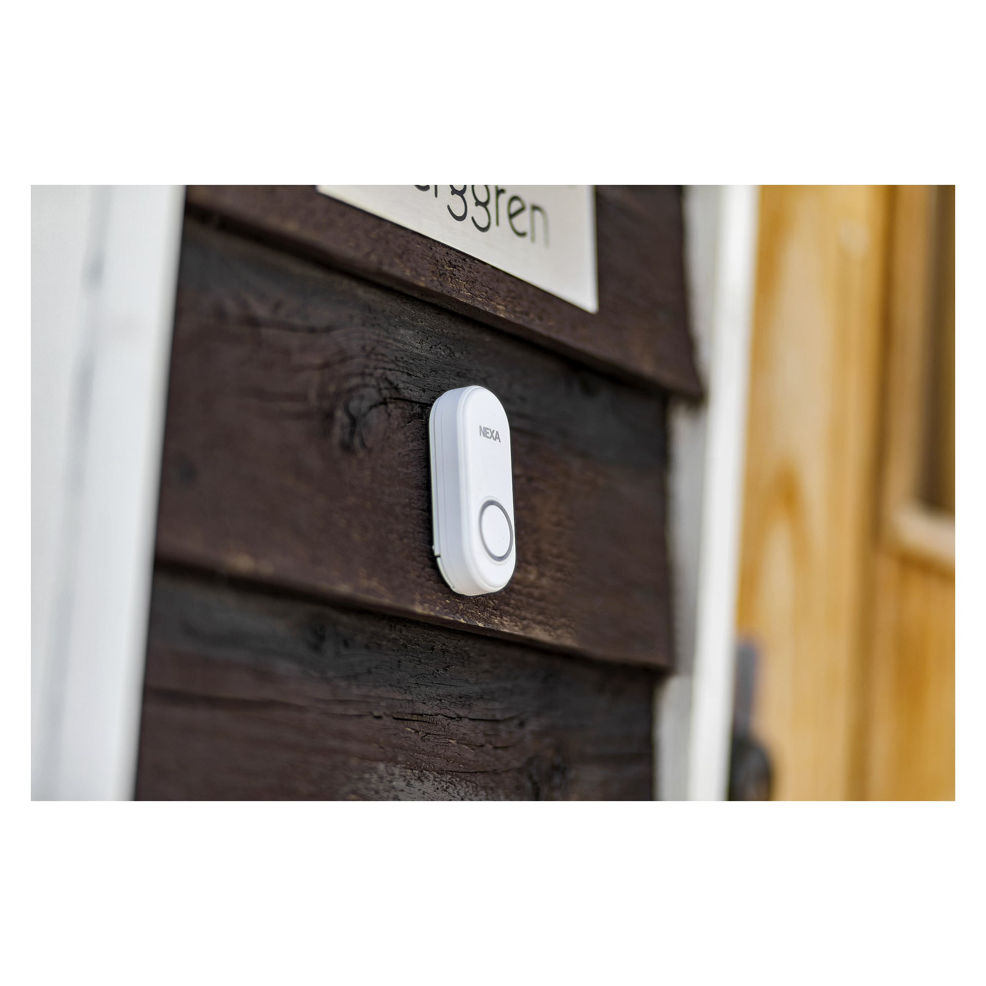 En vit Nexa-dörrklocksknapp är installerad på en mörk trävägg bredvid en ljusbrun dörr, med en del av en skylt som innehåller text synlig ovanför knappen.