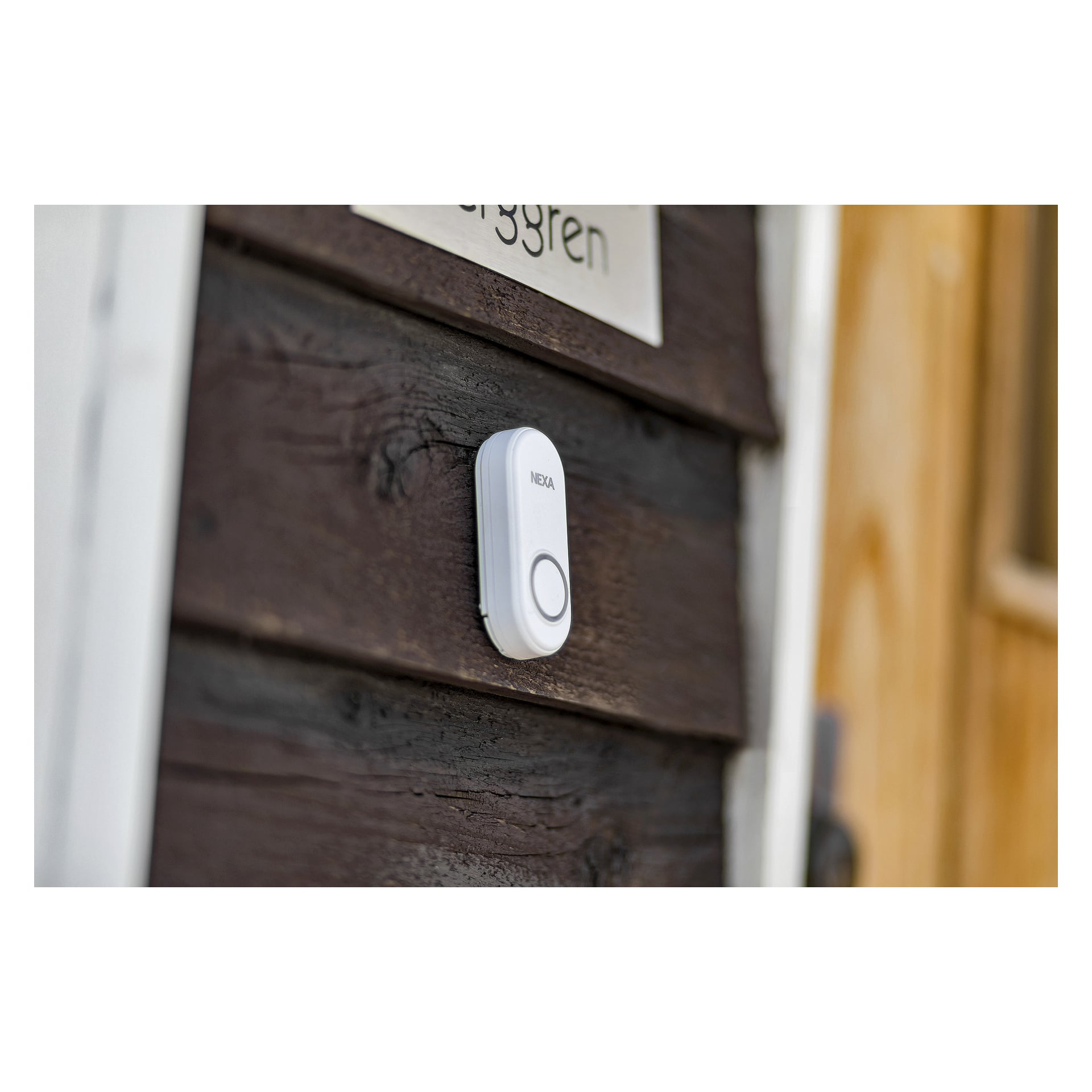 En vit Nexa Wireless dörrklocka MLR-1923 är installerad på en mörk trävägg bredvid en dörr, nära en namnskylt som bara är delvis synlig.