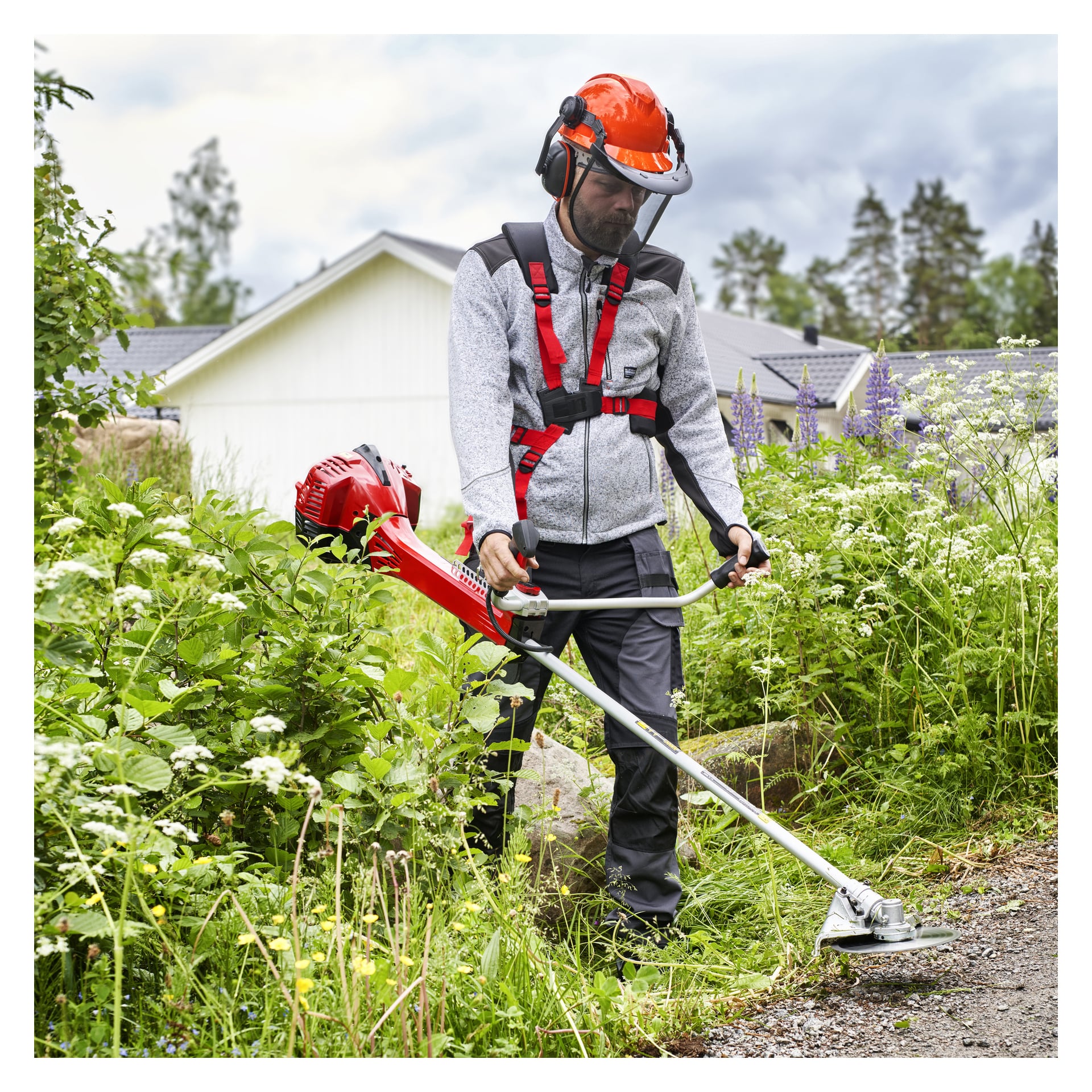 Med skyddsutrustning använder man en röd Meec Tools Brush-skärare för att rensa högt gräs och växter vid en trädgårdsgång, med hus och träd i fjärran.