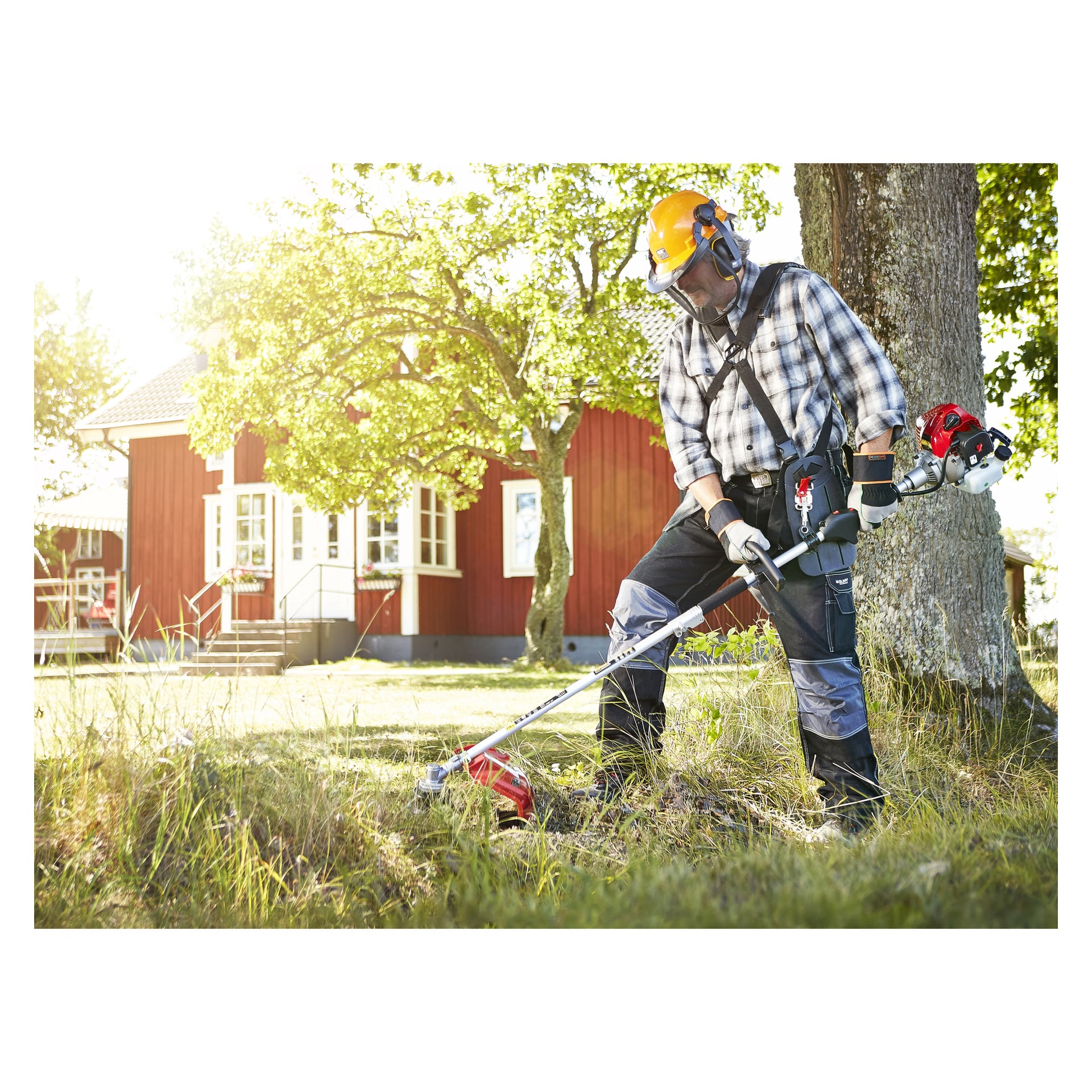 En person utrustad med säkerhetsutrustning använder en Meec Tools bensinsåg bredvid ett träd på en gräsmatta. Bakom dem står ett rött trähus med vita lister, upplyst av solljus.